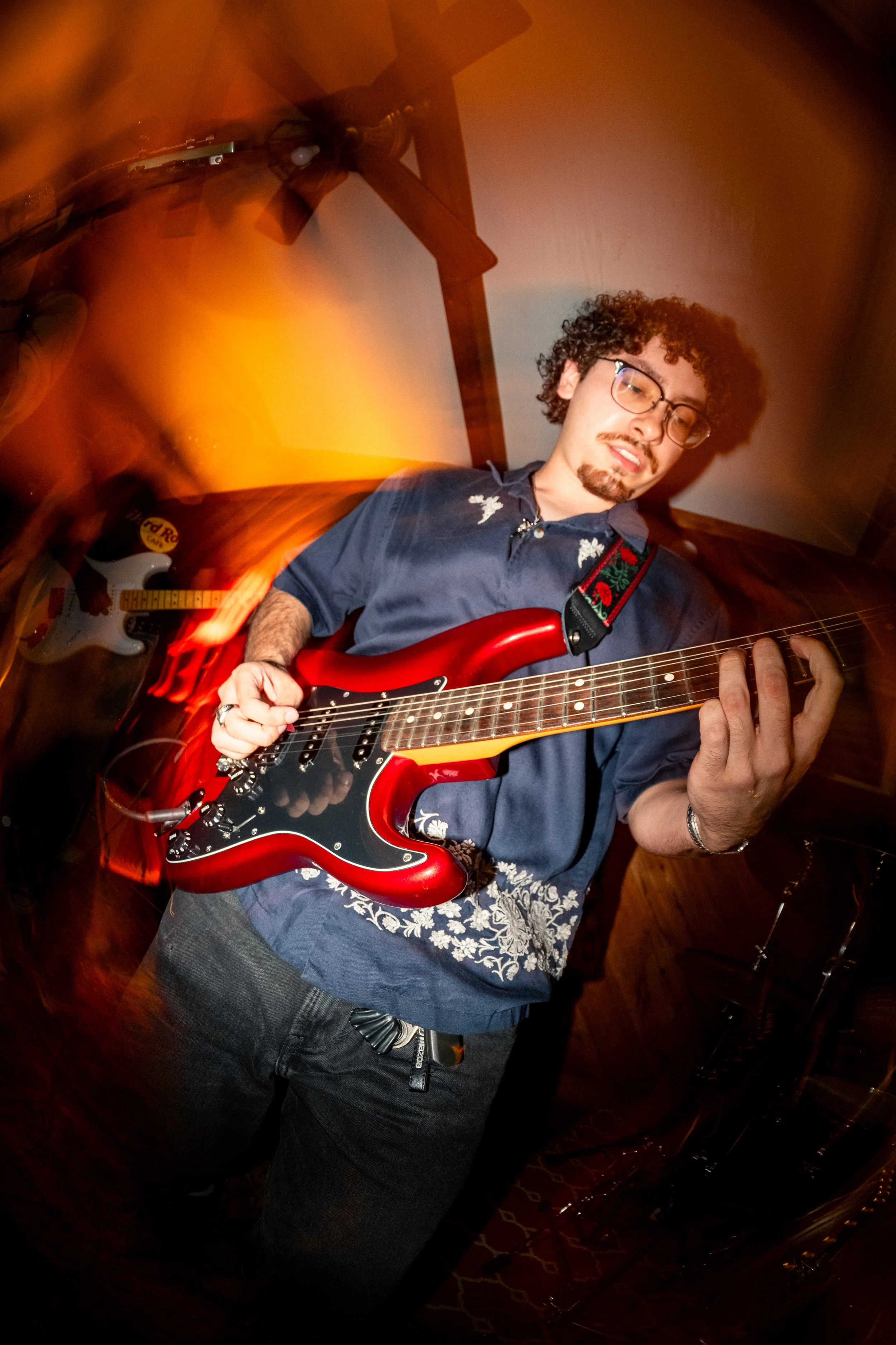 Man with curly hair and glasses playing a red electric guitar, wearing a navy shirt with floral embroidery, in a room with wooden details and yellow-orange lighting.