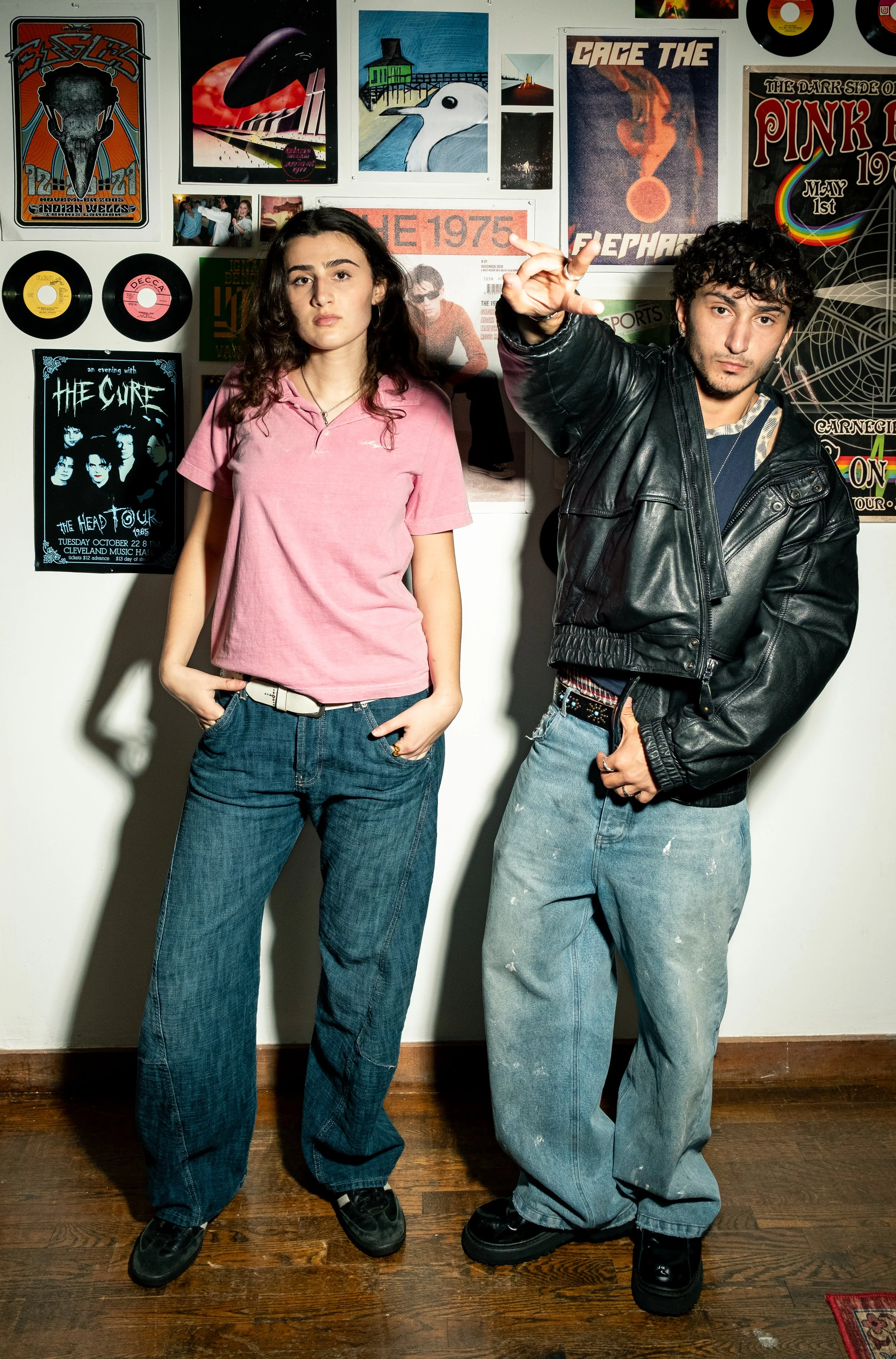 Two young adults standing in front of a wall decorated with music posters, vinyl records, and artwork. The woman on the left has long curly hair, wearing a pink polo shirt and loose jeans, with her hands in her pockets. The man on the right has dark 