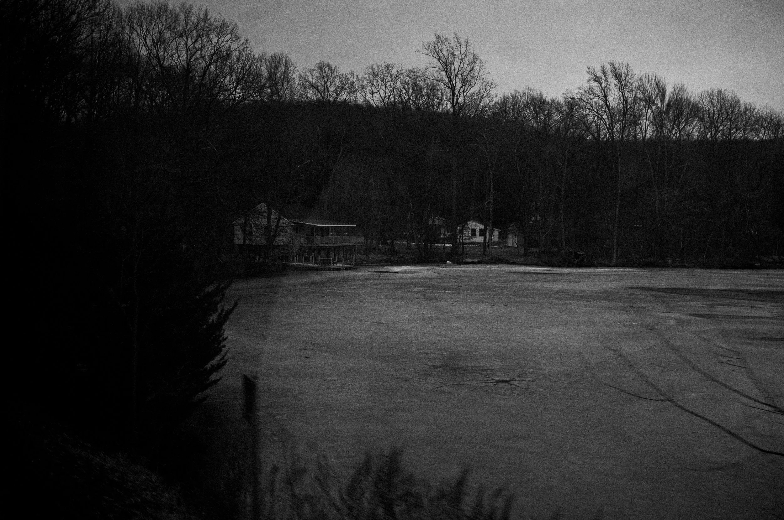 A black and white photo of a lake with a few houses in the background, surrounded by trees with no leaves, indicating it may be winter.