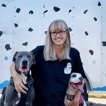 A woman with glasses and long blonde hair smiling, holding two dogs, standing in front of a white wall with blue and black abstract shapes.