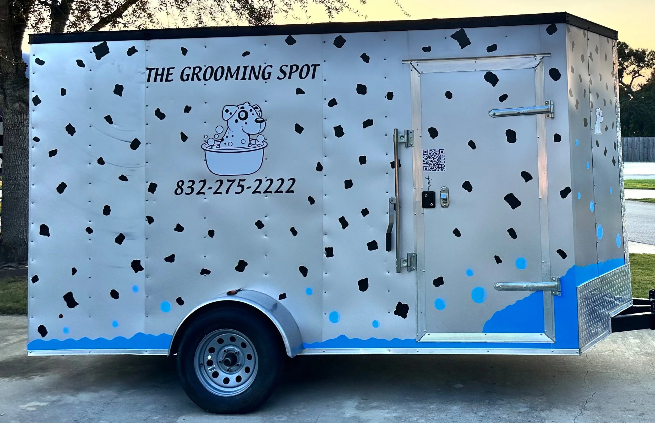 A gray food truck decorated with black and blue spots, featuring a cartoon dog in a bath, and the text 'The Grooming Spot' with a phone number.