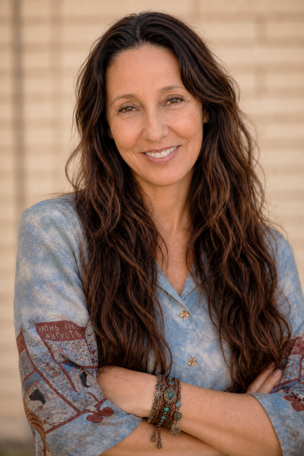 Alessandra, a woman with long wavy brown hair smiling, wearing a light blue patterned shirt and multiple bangles on her wrist, standing in front of a tan brick wall.