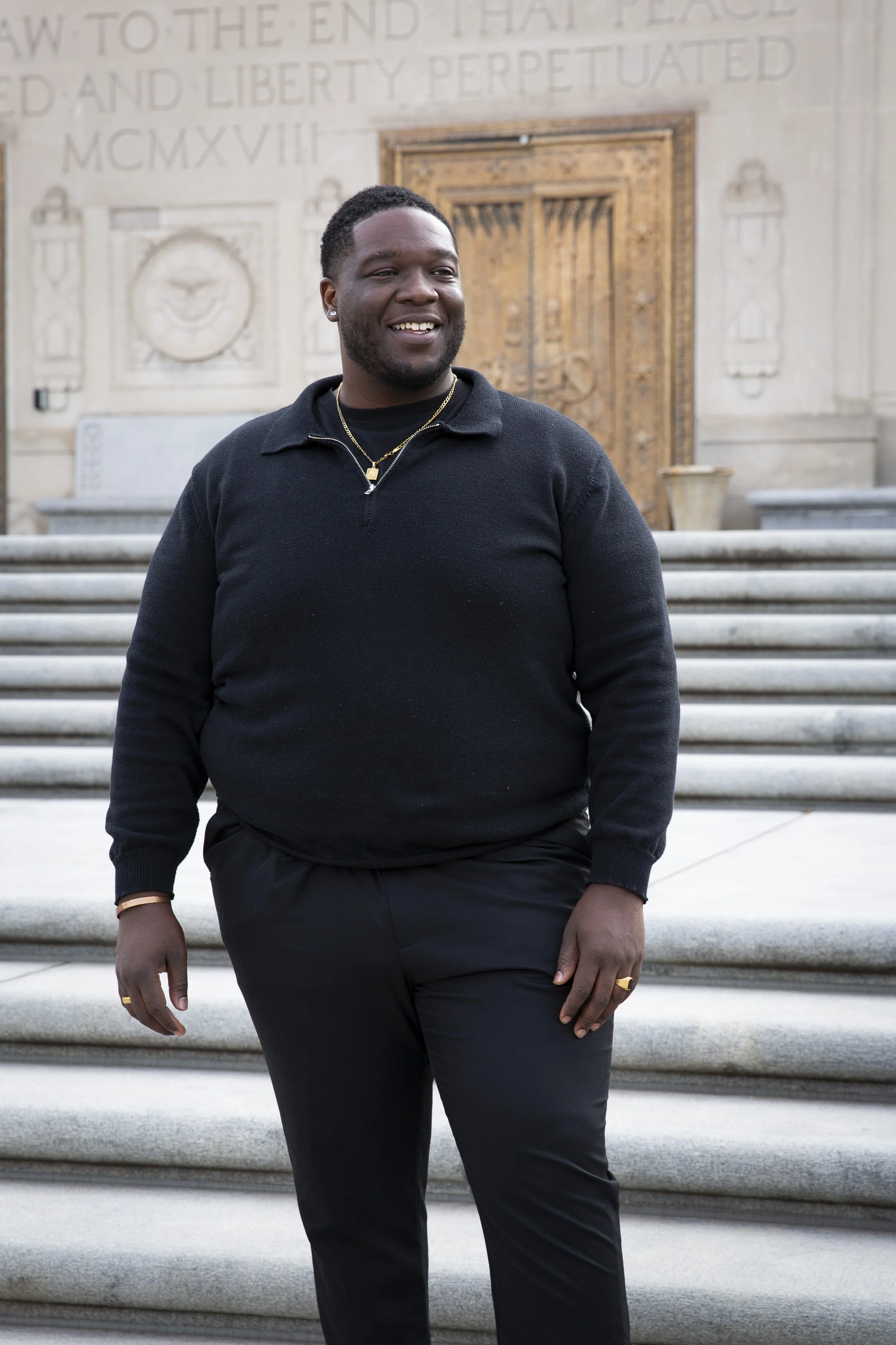 A smiling Black man in a black long-sleeve shirt and black pants standing on stairs in front of a stone building with engraved text and ornate doorway.