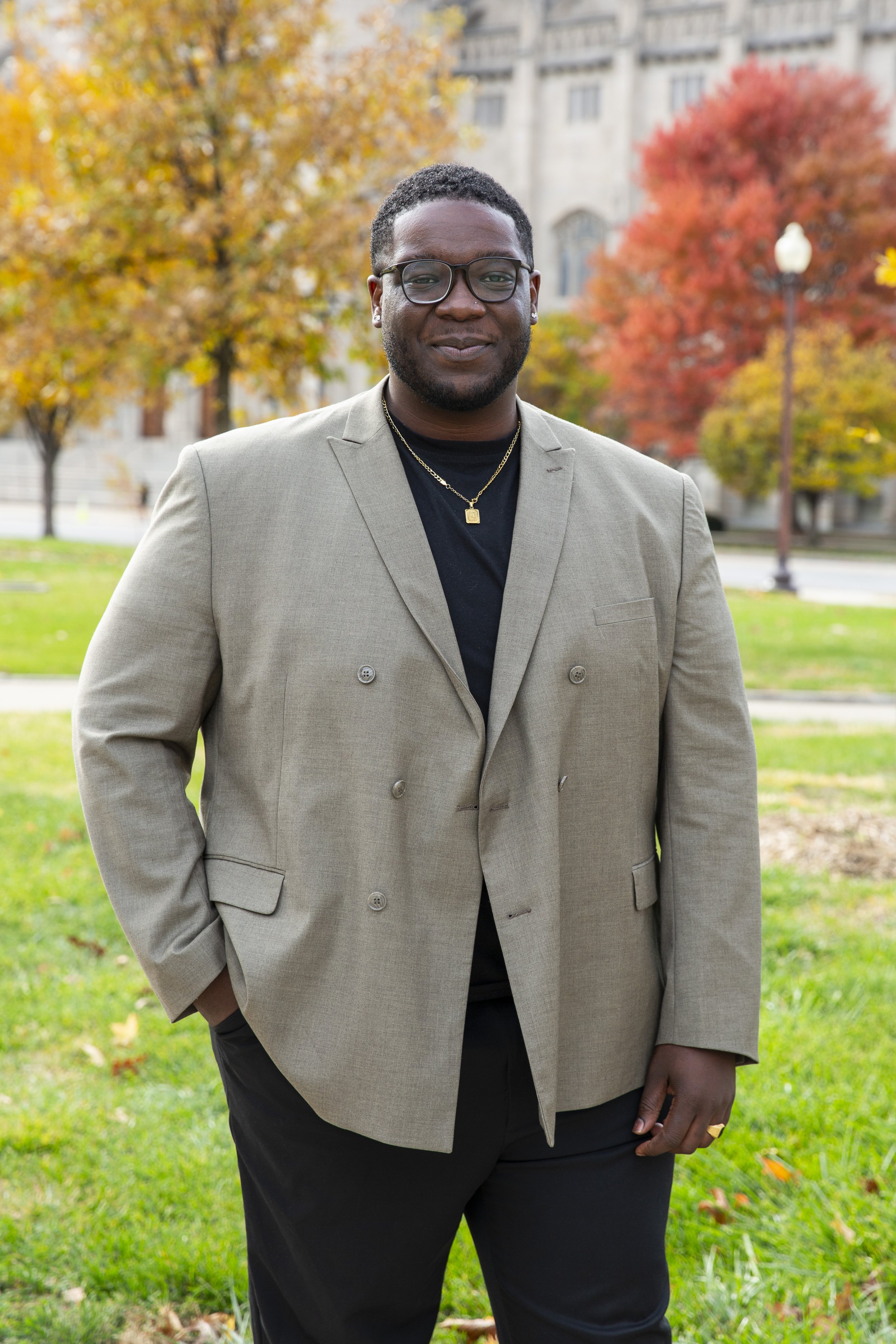 A man standing outdoors in a park during fall, wearing a beige blazer over a black shirt, glasses, and jewelry with trees showing autumn colors in the background.