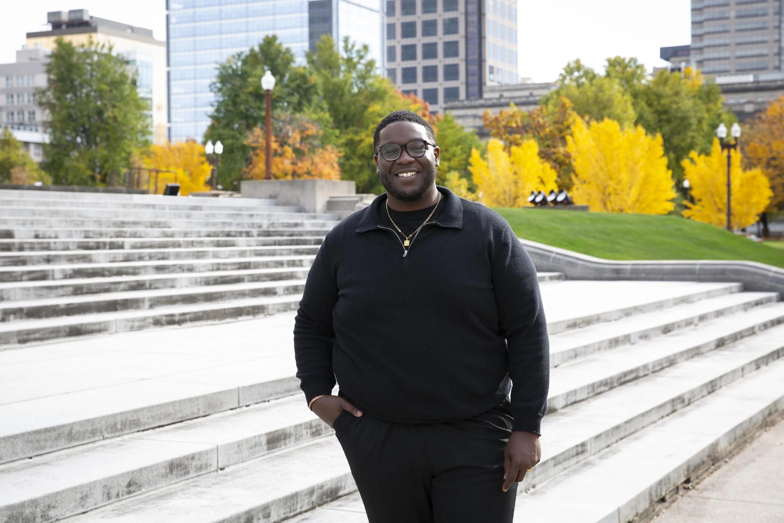 A smiling man wearing glasses and a black sweater standing outdoors near steps and trees with yellow and orange leaves in an urban park.