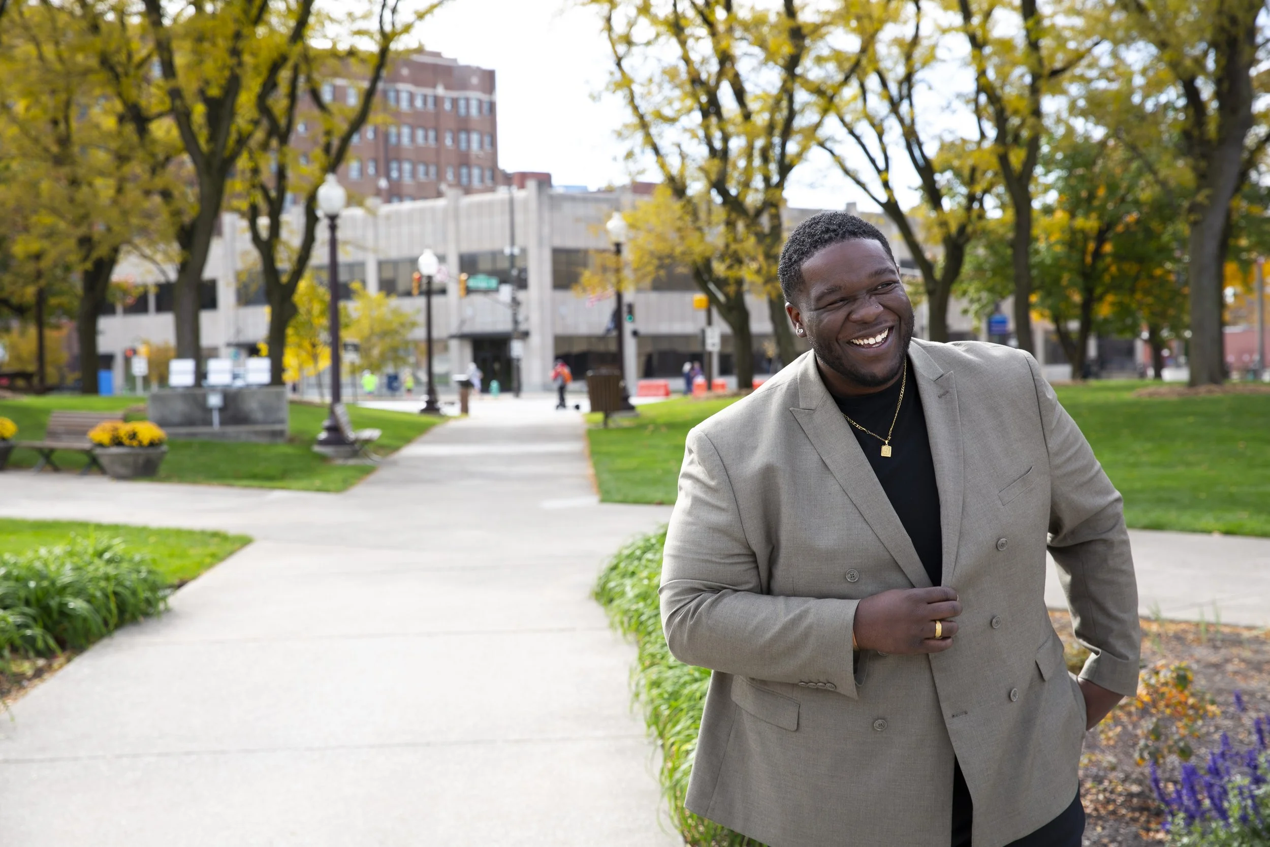 A cheerful man in a beige blazer and black shirt, smiling as he walks in a park with green grass, colorful flowers, and tall trees with autumnal leaves. Buildings are visible in the background.