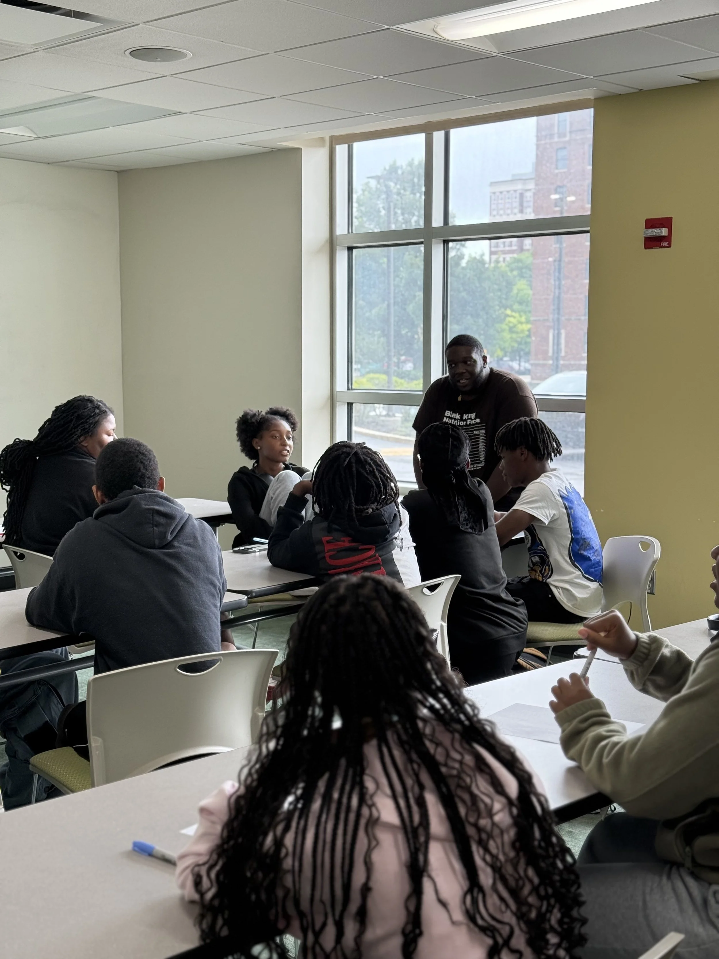 A classroom with students sitting at tables and a man standing near a window, engaging with the students during a lesson.