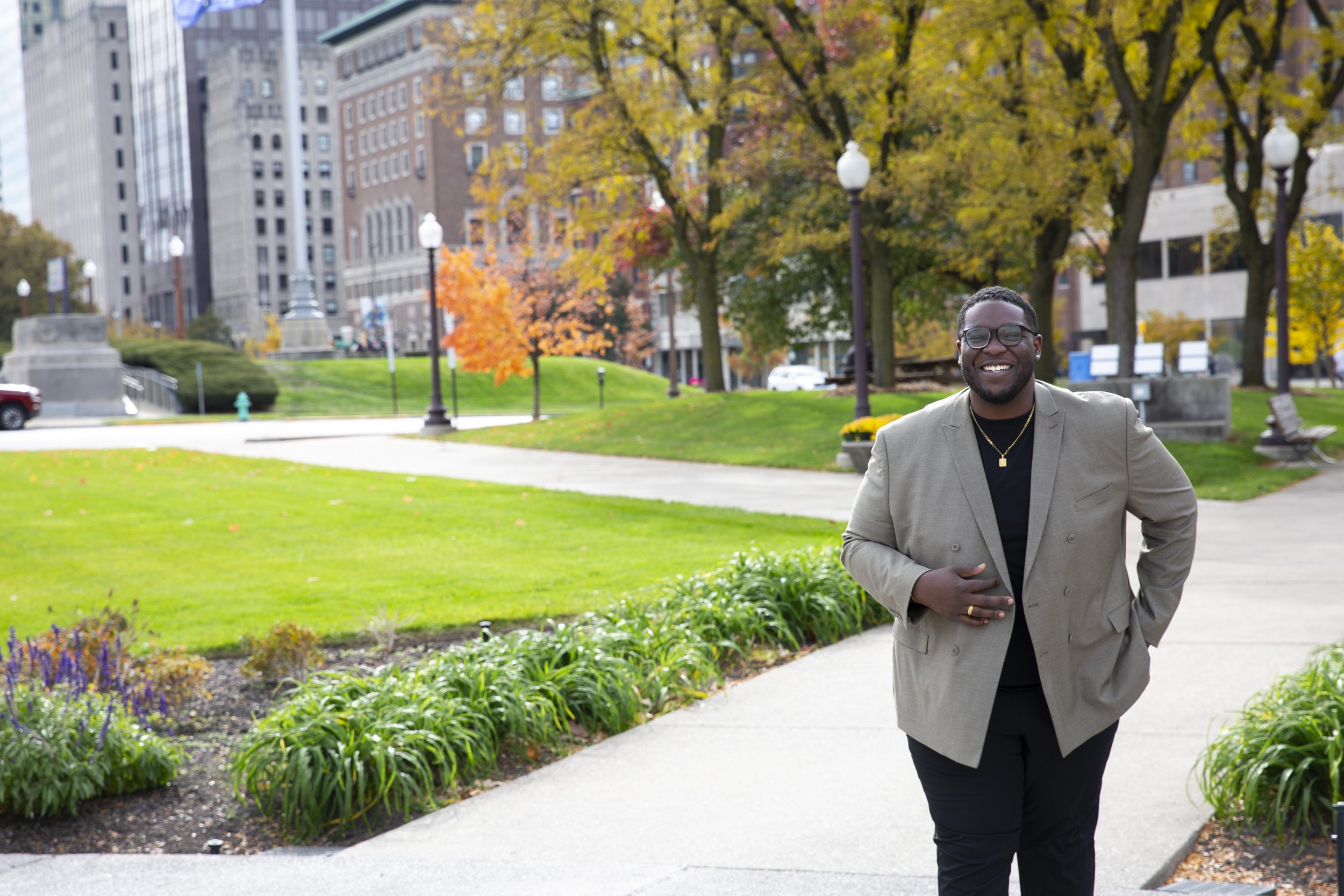 A smiling man wearing glasses, a tan blazer, and black pants walking in a park with autumn foliage and city buildings in the background.