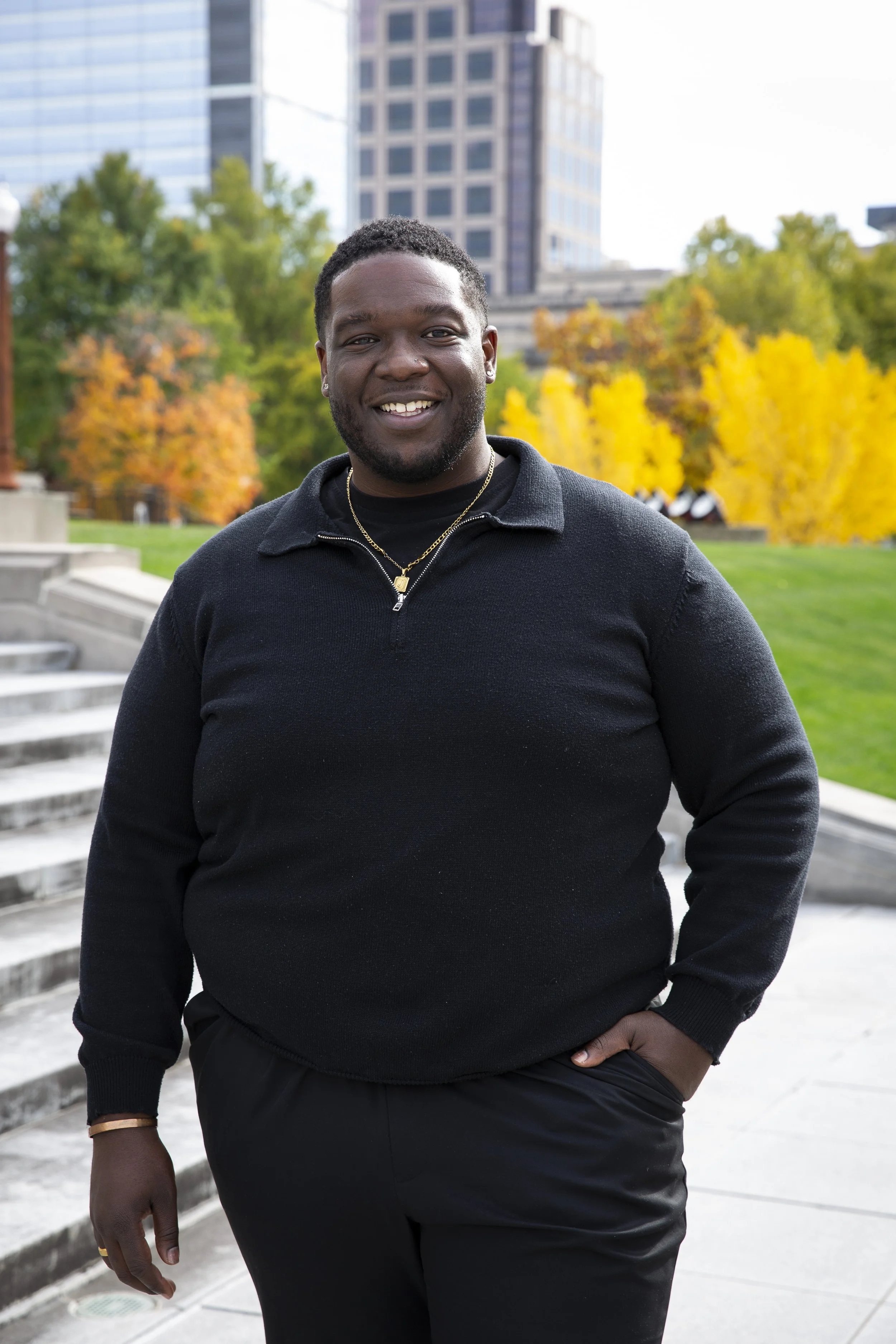 A smiling Black man wearing a black sweater and pants outdoors in an urban park with autumn trees and buildings in the background.