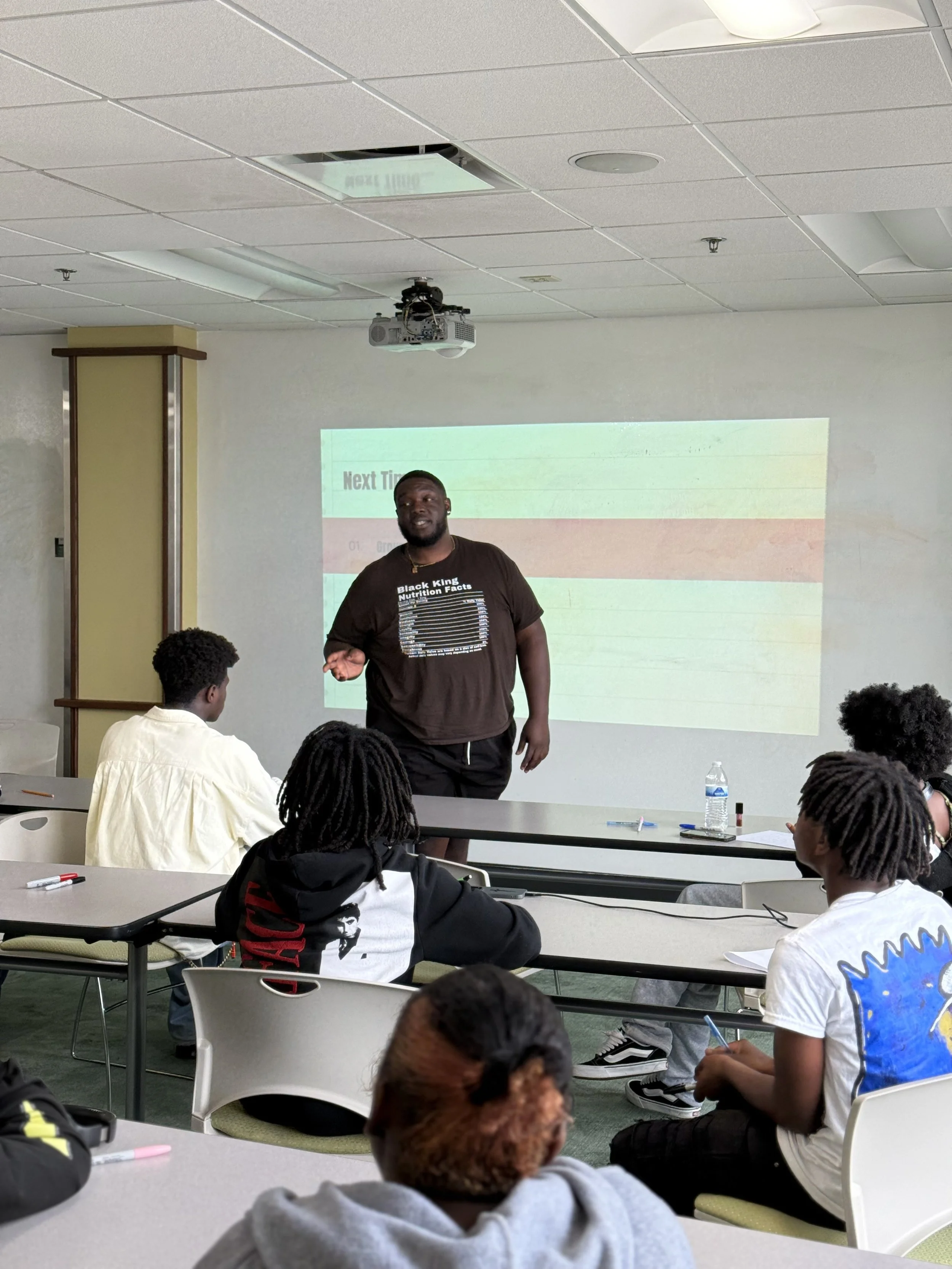 A man standing in front of a classroom with a projected presentation, engaging with students seated at tables, some taking notes and facing him.