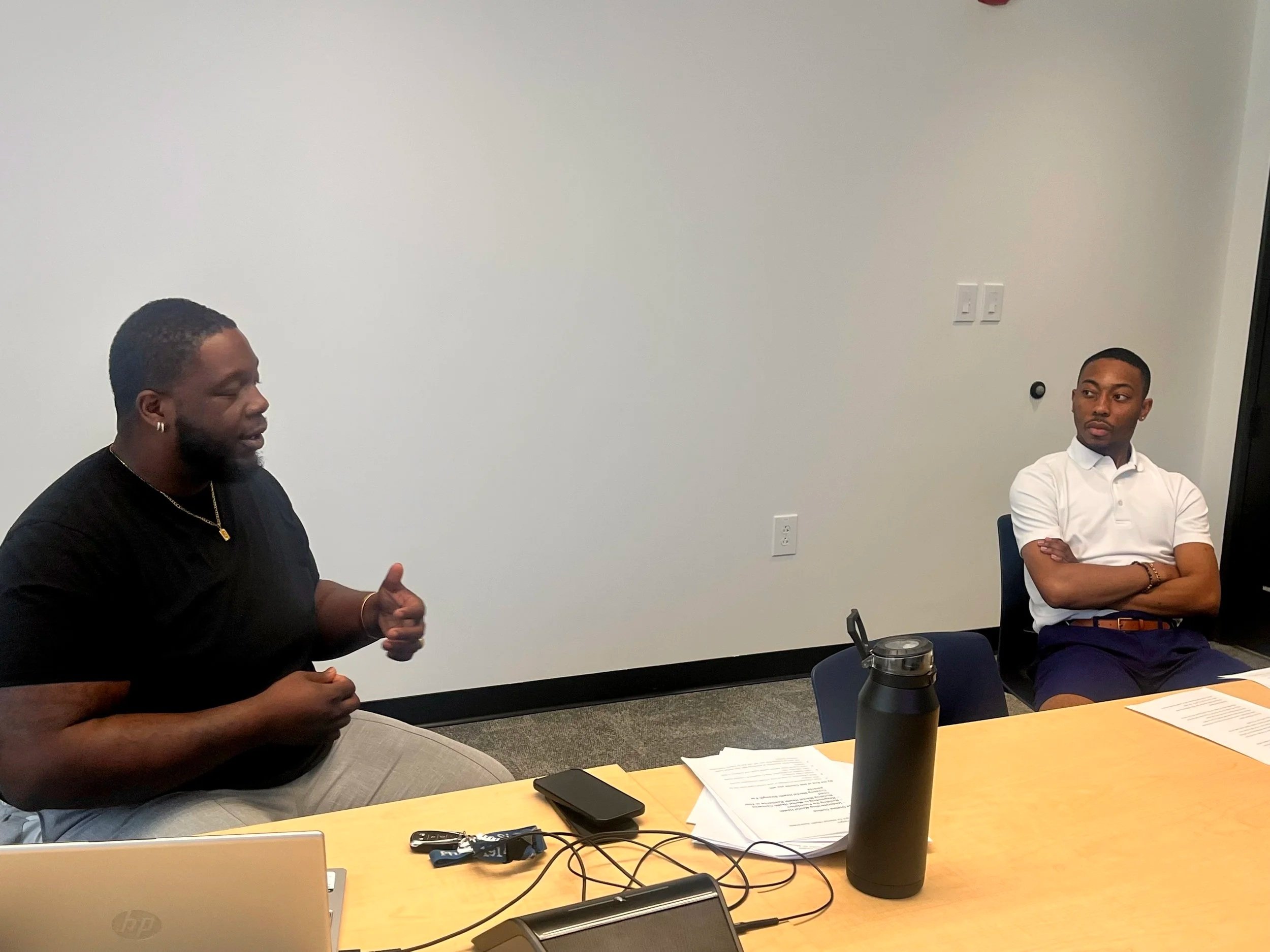 Two men sitting at a conference table engaged in a discussion, with papers, a laptop, a water bottle, and electronic devices on the table.