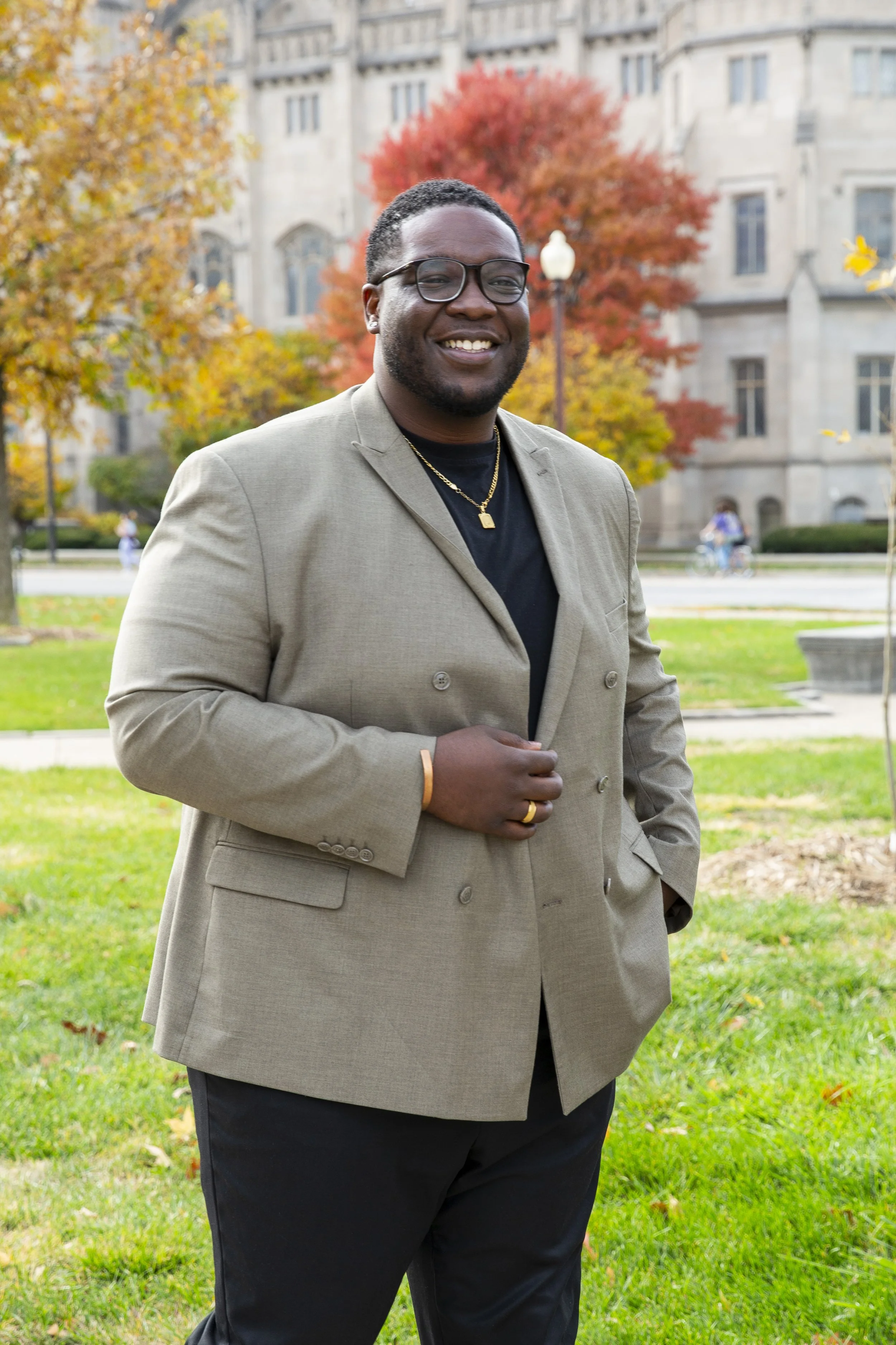A smiling man wearing glasses, a beige blazer, black shirt, and gold jewelry standing outdoors in front of autumn trees and a historic building.