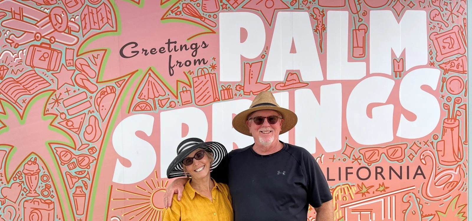A smiling couple are standing in front of a large pink mural that says "Greetings from Palm Springs" with images of desert resort foods and other vacation items. They are both wearing sunglasses with broad brim hats.