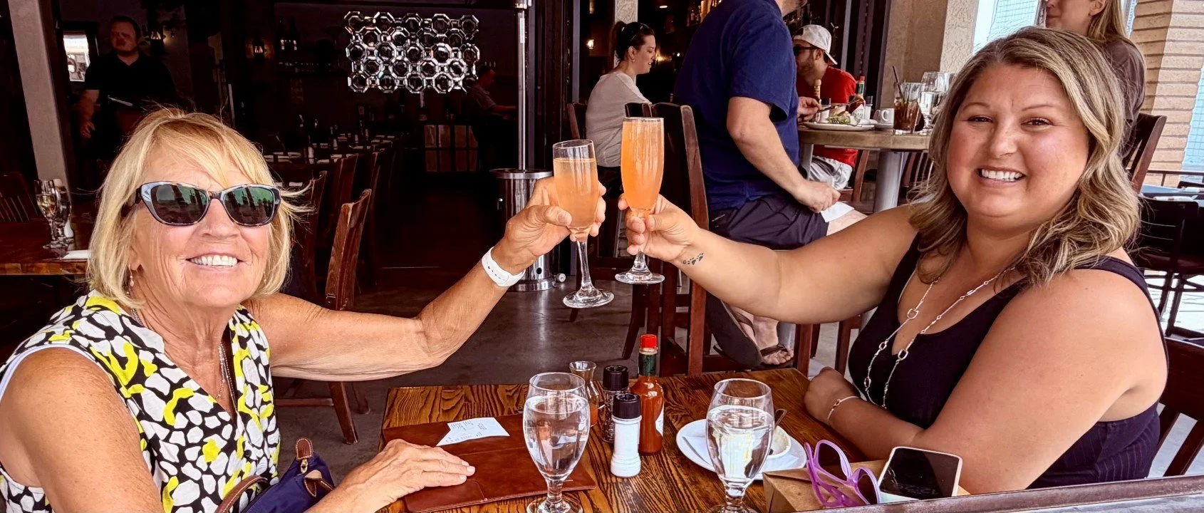 Two women with raised champagne glasses making a toast sitting on a restaurant patio area smiling and looking happy with the one on the left wearing sunglasses.