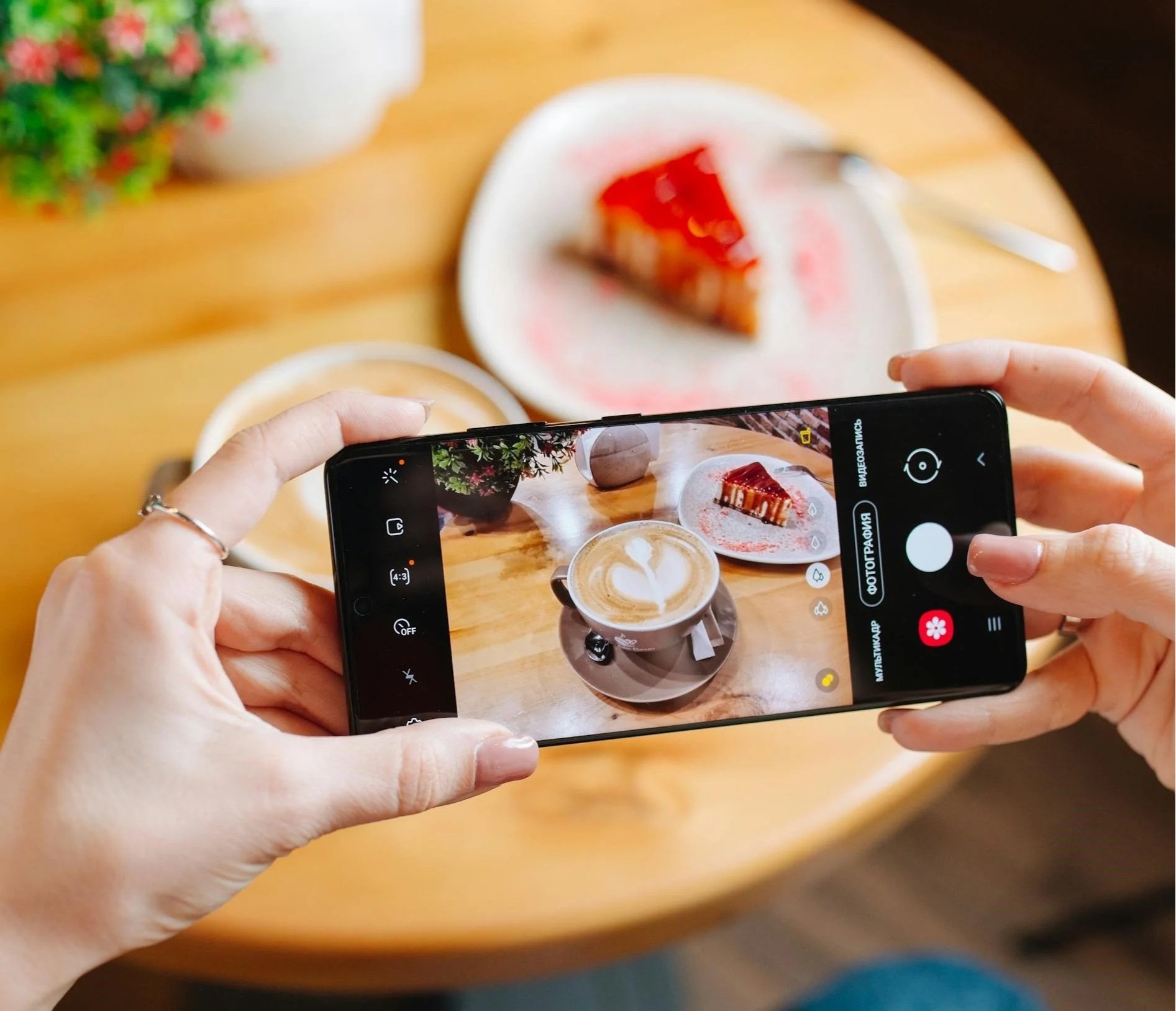 Person taking a photo of a coffee and a slice of cake on a wooden table with a smartphone.