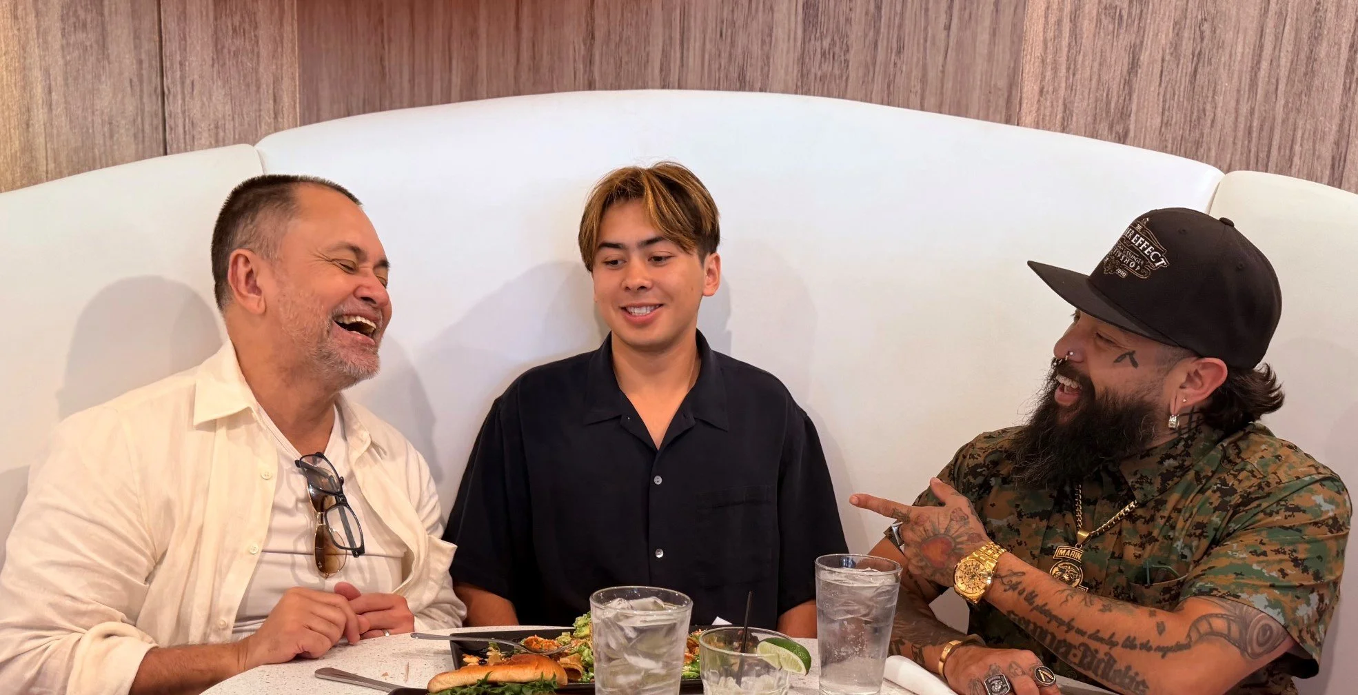 Three men sitting in a restaurant booth smiling and laughing with a wood paneled wall and the white cushion back of the booth behind them. One man is heavily tattooed and wearing lots of jewelery.
