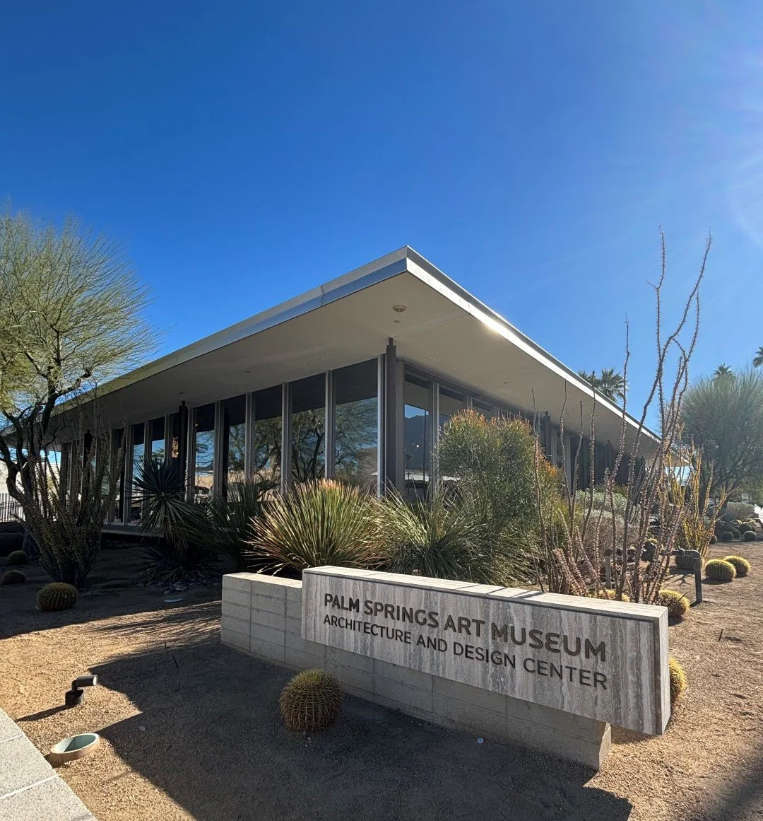 Exterior view of Palm Springs Art Museum Architecture and Design Center  showing the sign and building under a clear blue sky with cacti and dark shadows cast by the building and concrete block sign. sign