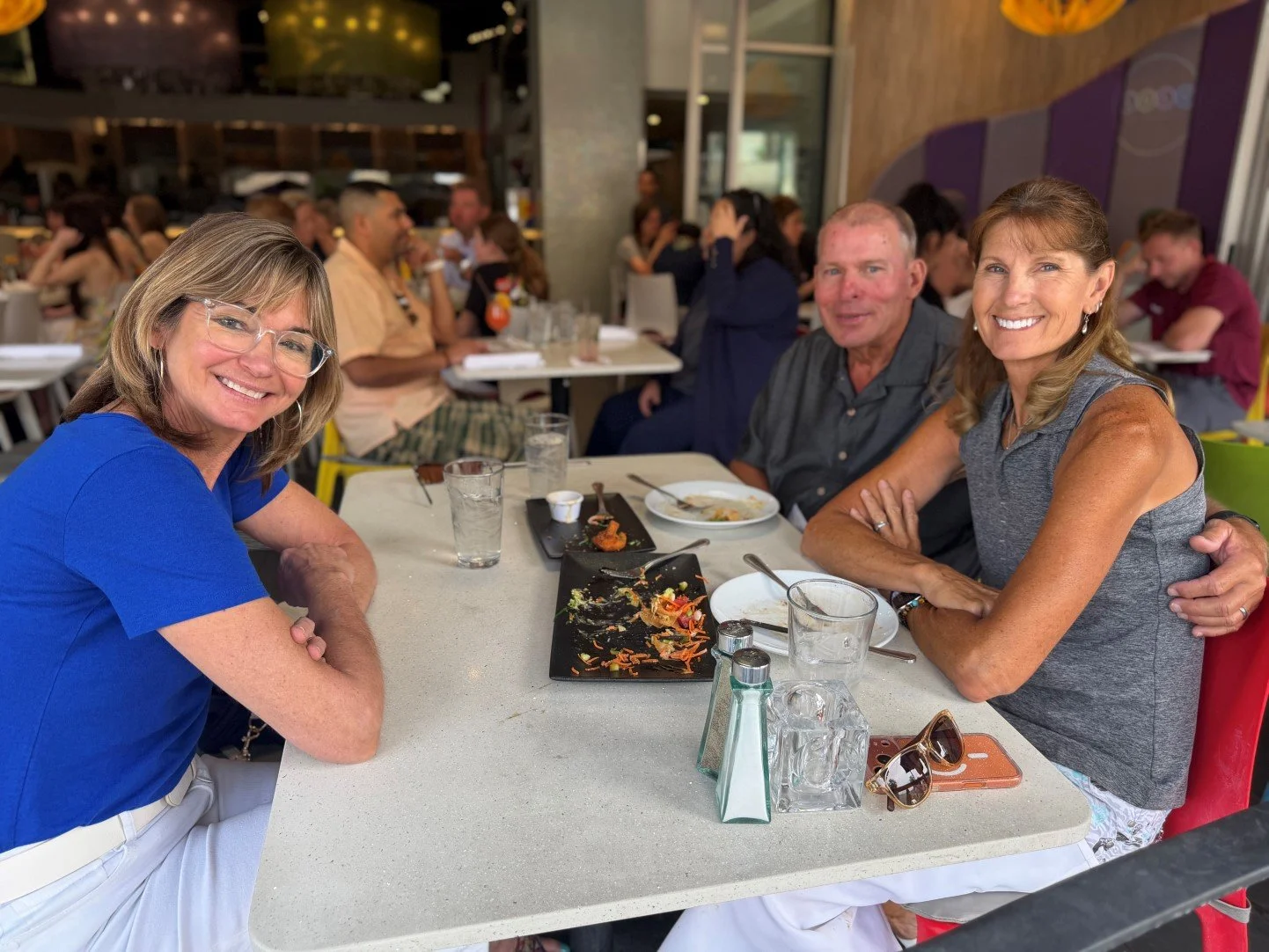A group of three happy people sitting in an open air restaurant who just finished two appetizer plates and are looking at the photographer with big smiles on their faces