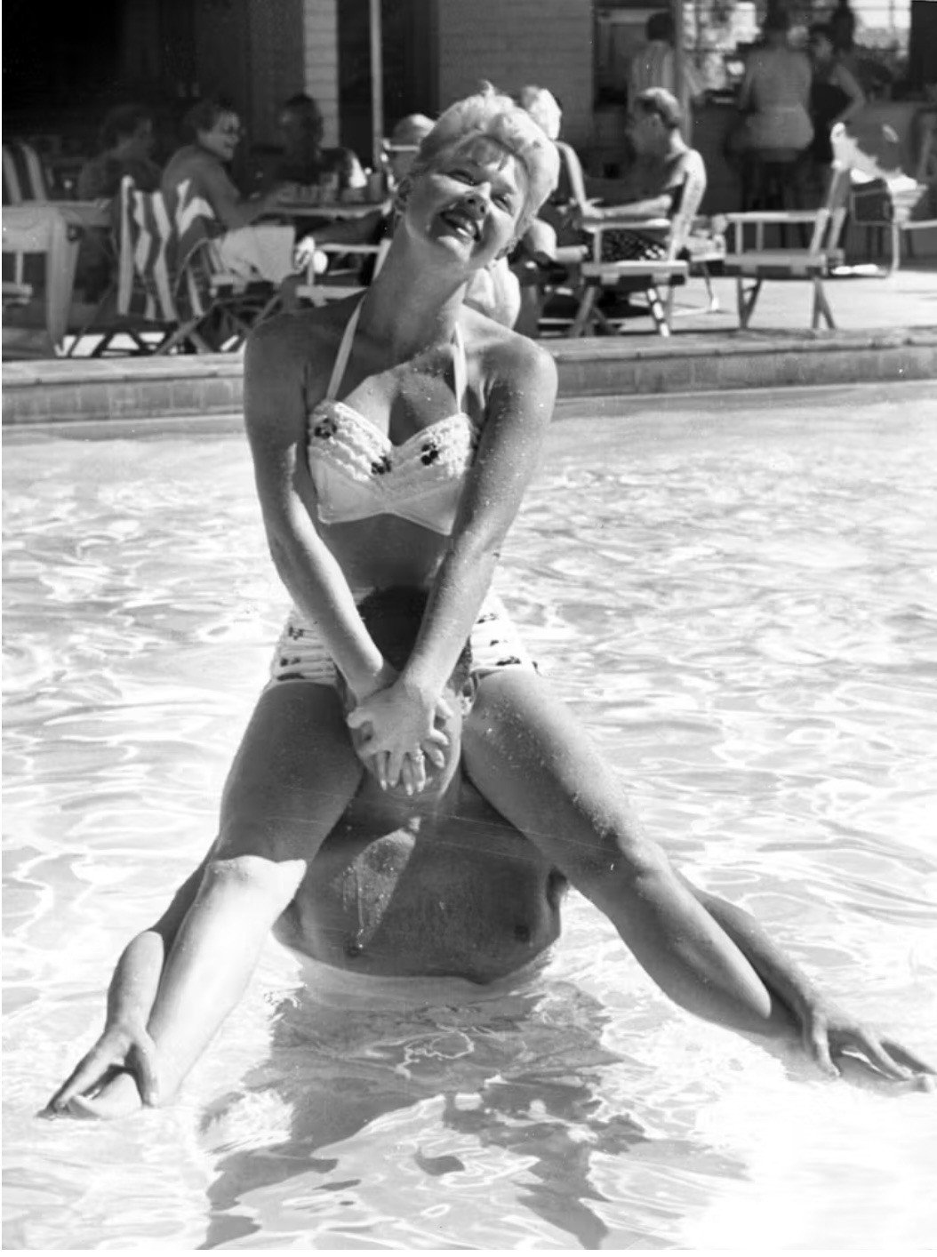 A woman in a swimsuit is sitting on a man's shoulders in a pool, smiling at the camera. People are sitting at tables in the background.