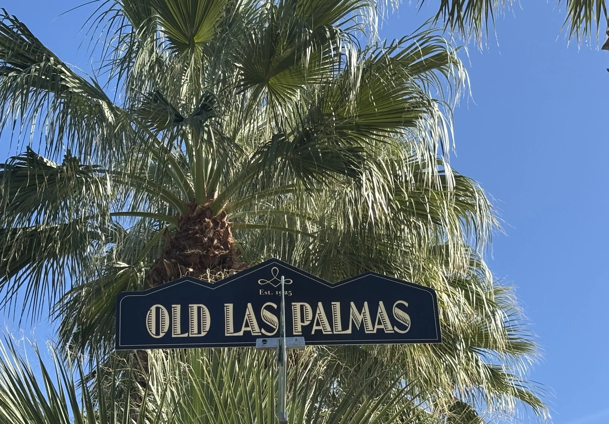 A street sign with the words "Old Las Palmas" is in front of palm tree fronds wiht a bright blue sky in the background.