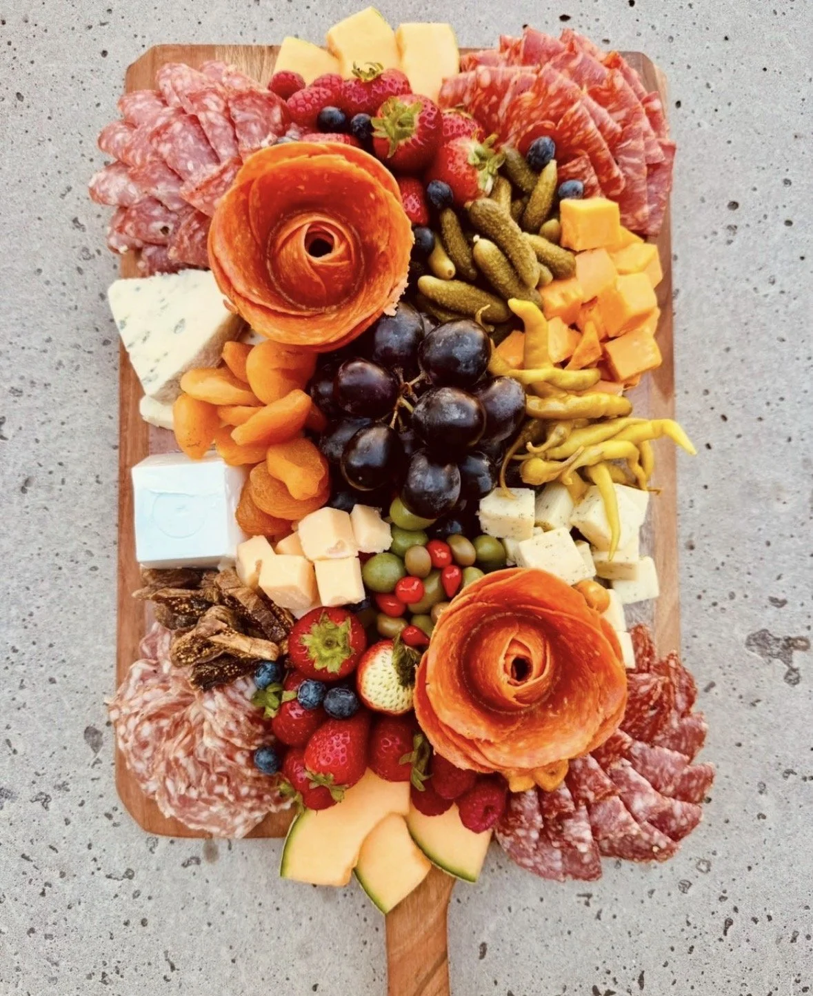 Artisan quality deli meats and cheeses on a board with fresh and dried fruit, olives, pickles and peppers. The food is attractively displayed with two of the sliced meats arranged in a flower design.