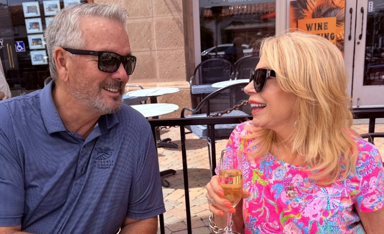 A man and woman looking at each other while sitting at an outdoor cafe, smiling and talking, with the woman holding a glass of champagne, both wearing sunglasses, and there are tables and chairs behind them.