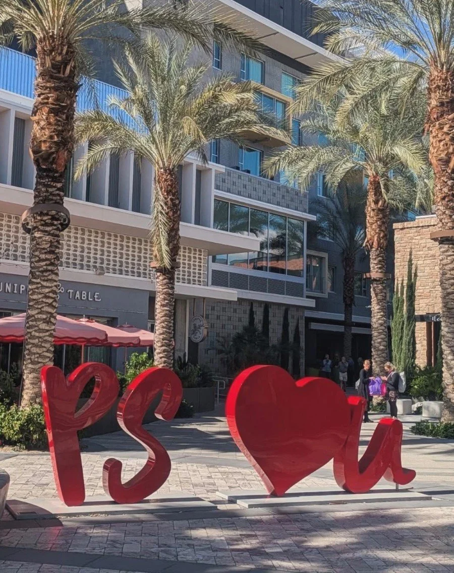 Large red metal sculpture spelling 'PS' '❤' 'u' in an outdoor plaza surrounded by palm trees and modern buildings with glass windows in downtown Palm Springs California.