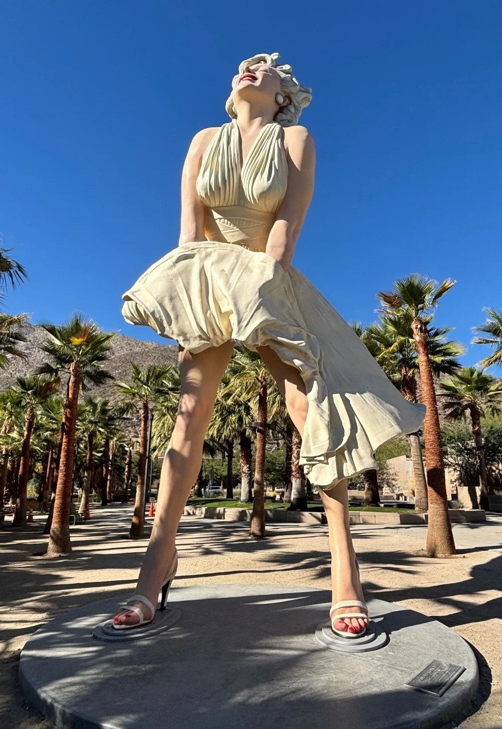 Large statue of Marilyn Monroe in a glamorous dress with high heels, standing outdoors with palm trees and mountains in the background under a clear blue sky in Palm Springs, California.