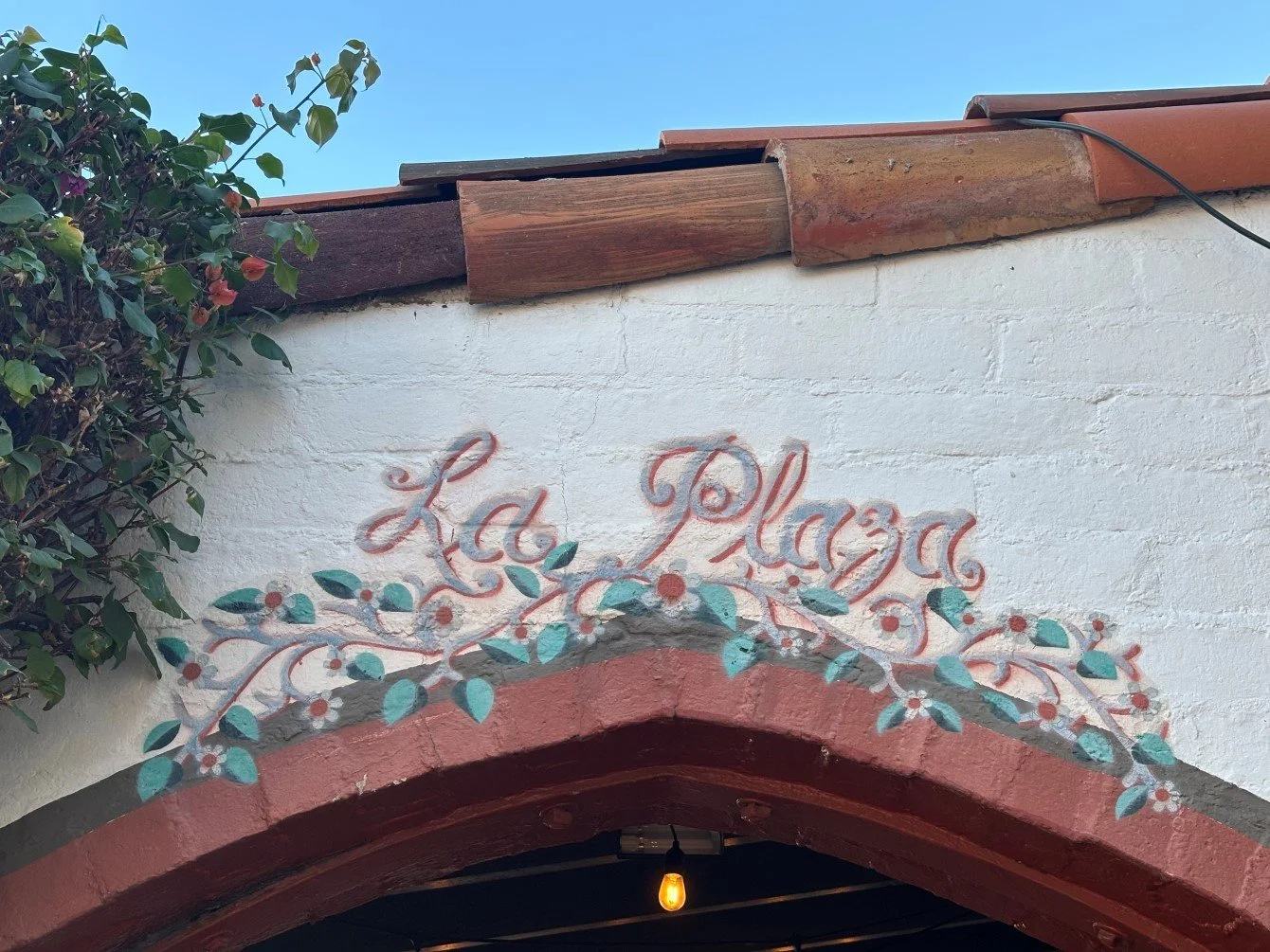 Storefront with a red tile roof with the white wall above a red brick Spanish style archway with the sign "La Plaza" written in red cursive above a painted green floral design on the white wall