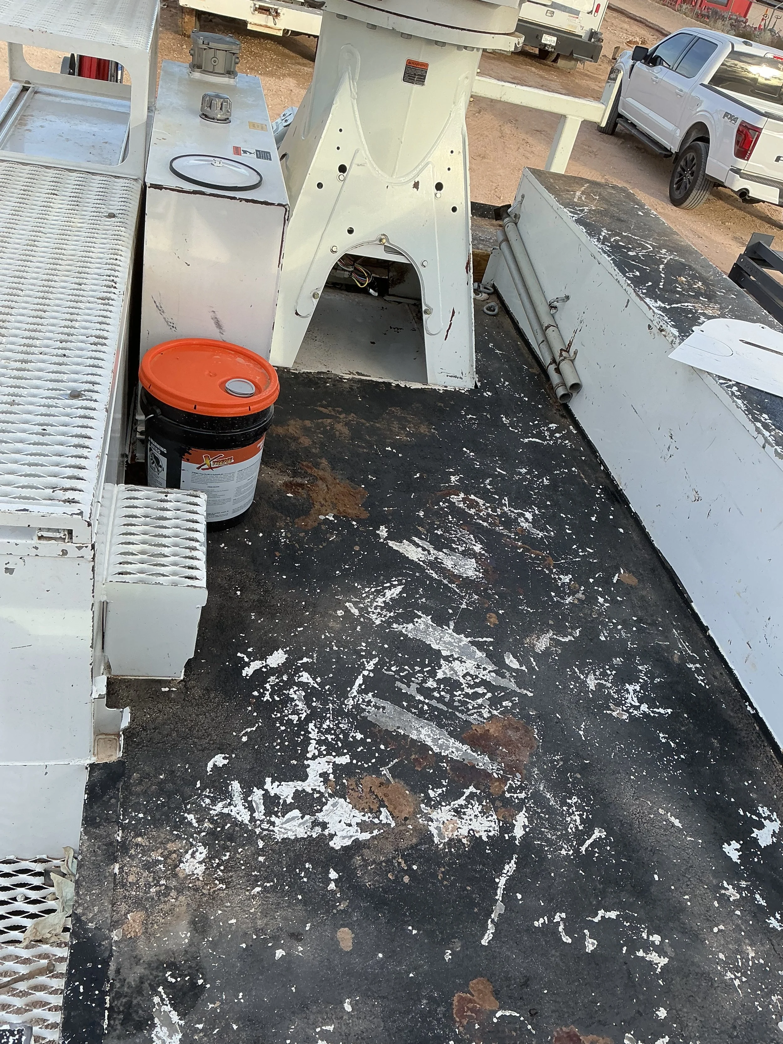 View of a metal work platform with rust and paint splatters, a bucket, and some machinery, with trucks parked in the background.