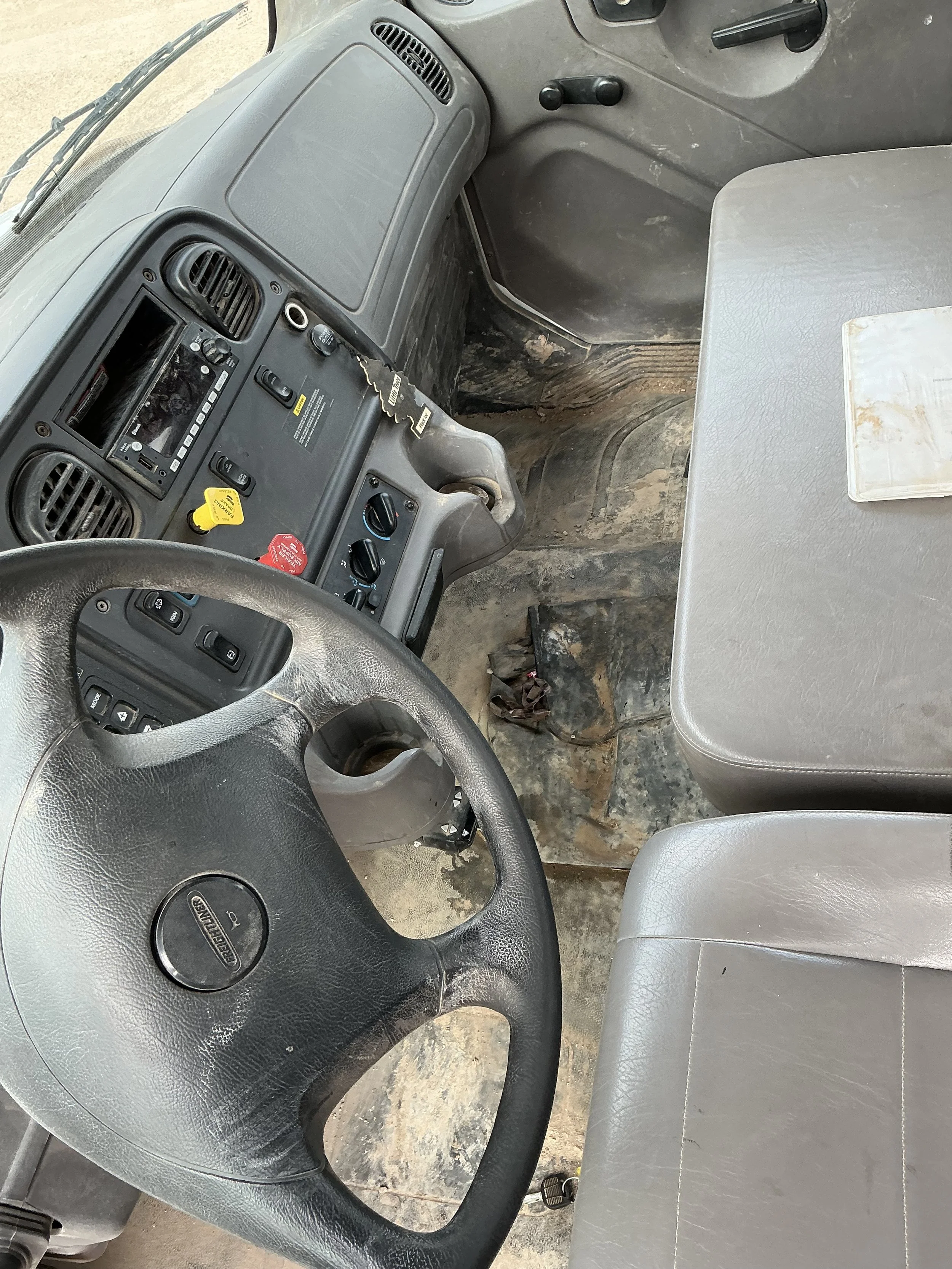 Interior of a work truck with a gray dashboard, steering wheel, and bench seat. The driver's side floor shows dirt and wear, with visible keys hanging from the ignition and a blank clipboard on the passenger seat.