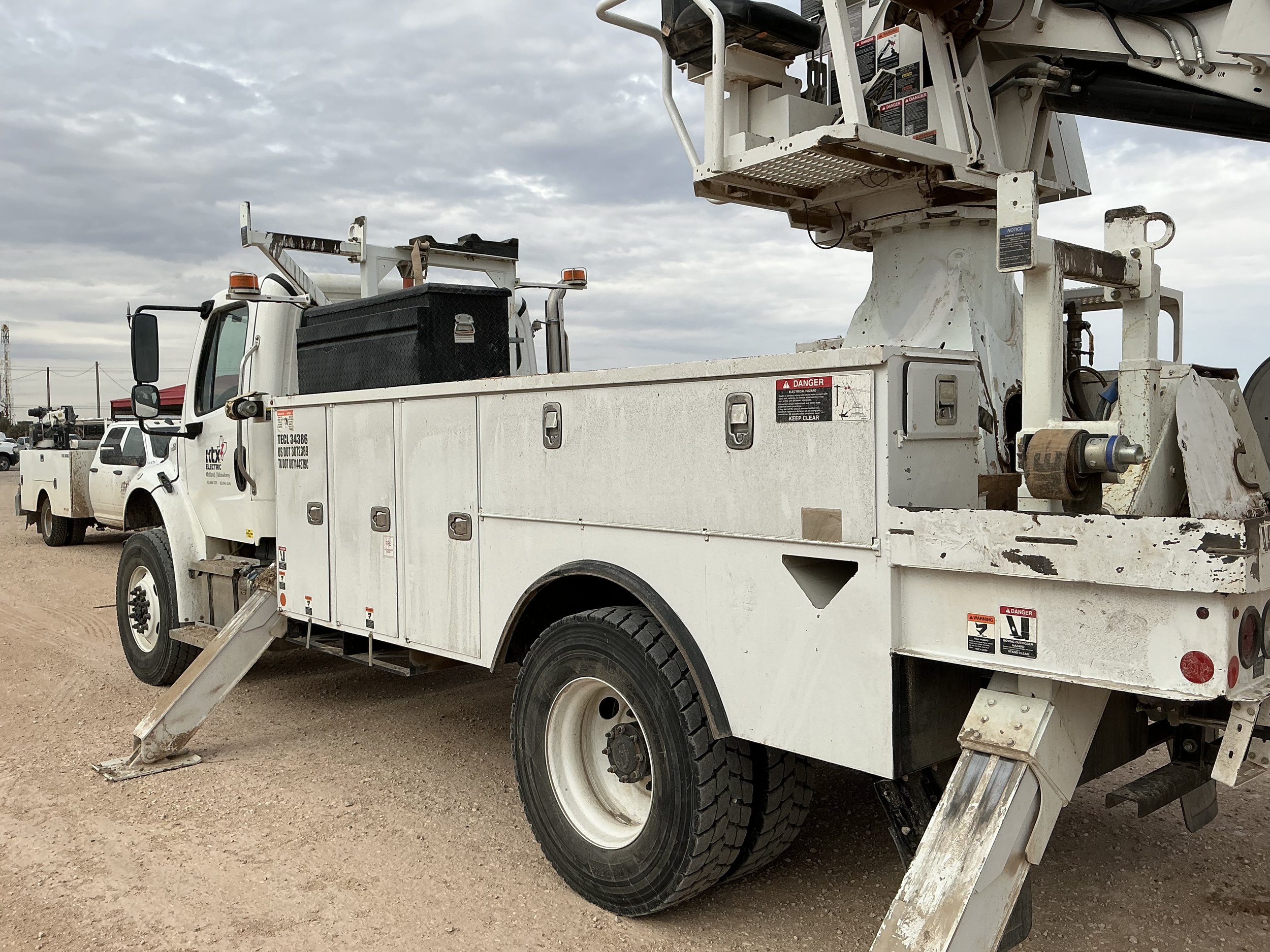 A white utility truck with an elevated work platform, parked on a dirt lot with other service vehicles in the background, under a cloudy sky.