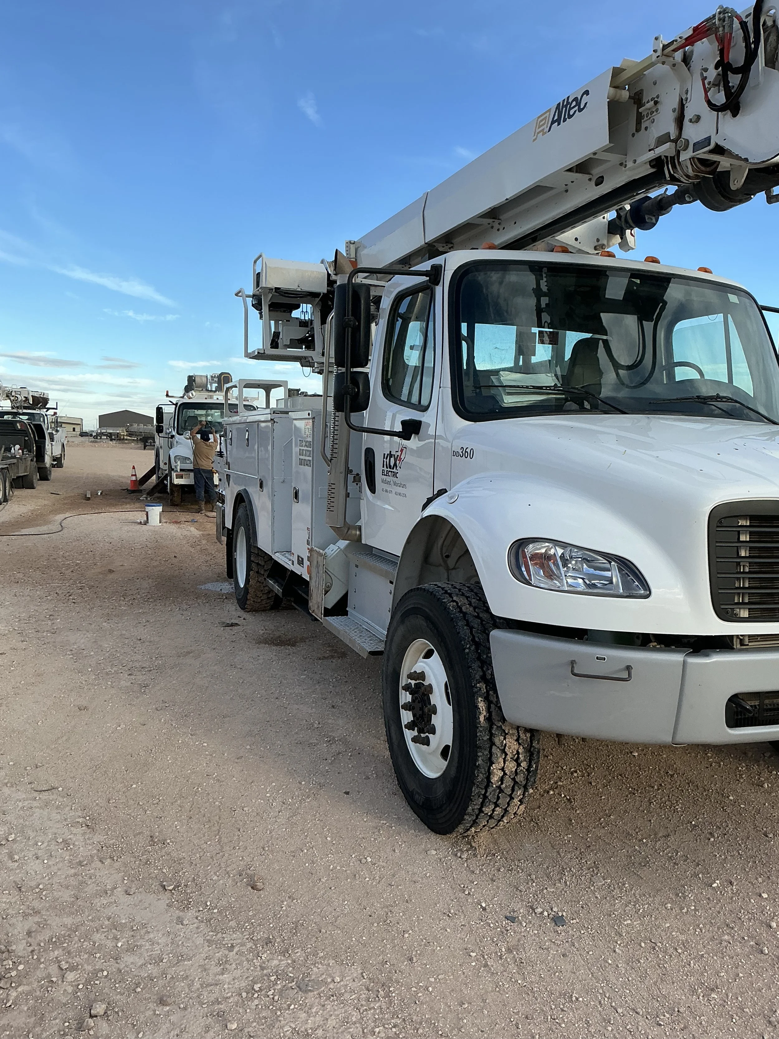 A white utility truck with a crane on top parked on a dirt lot, with a person working nearby and other trucks in the background under a blue sky.