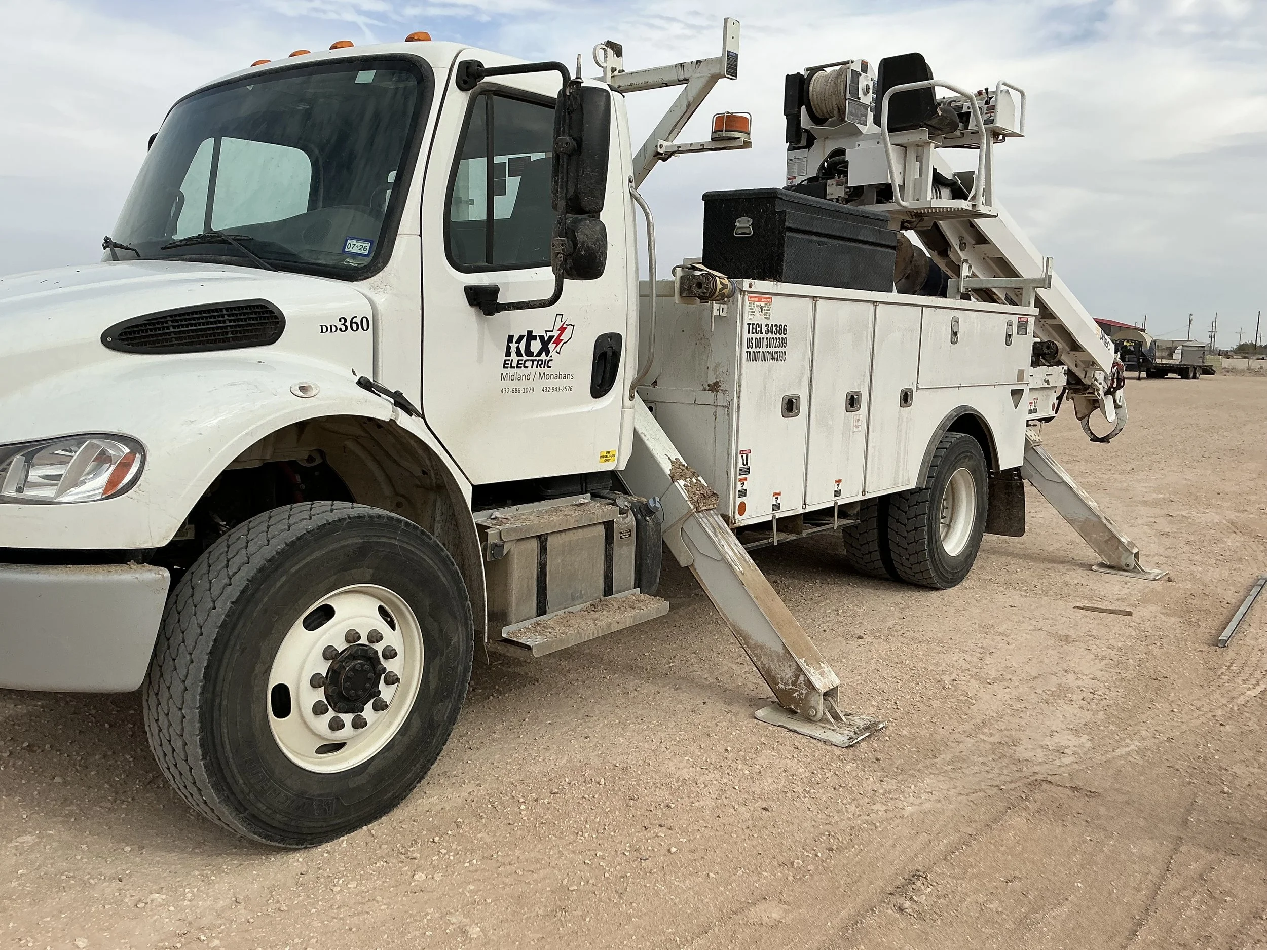 White utility truck with equipment, stabilizers, and a bucket lift, parked on a gravel lot under an overcast sky.