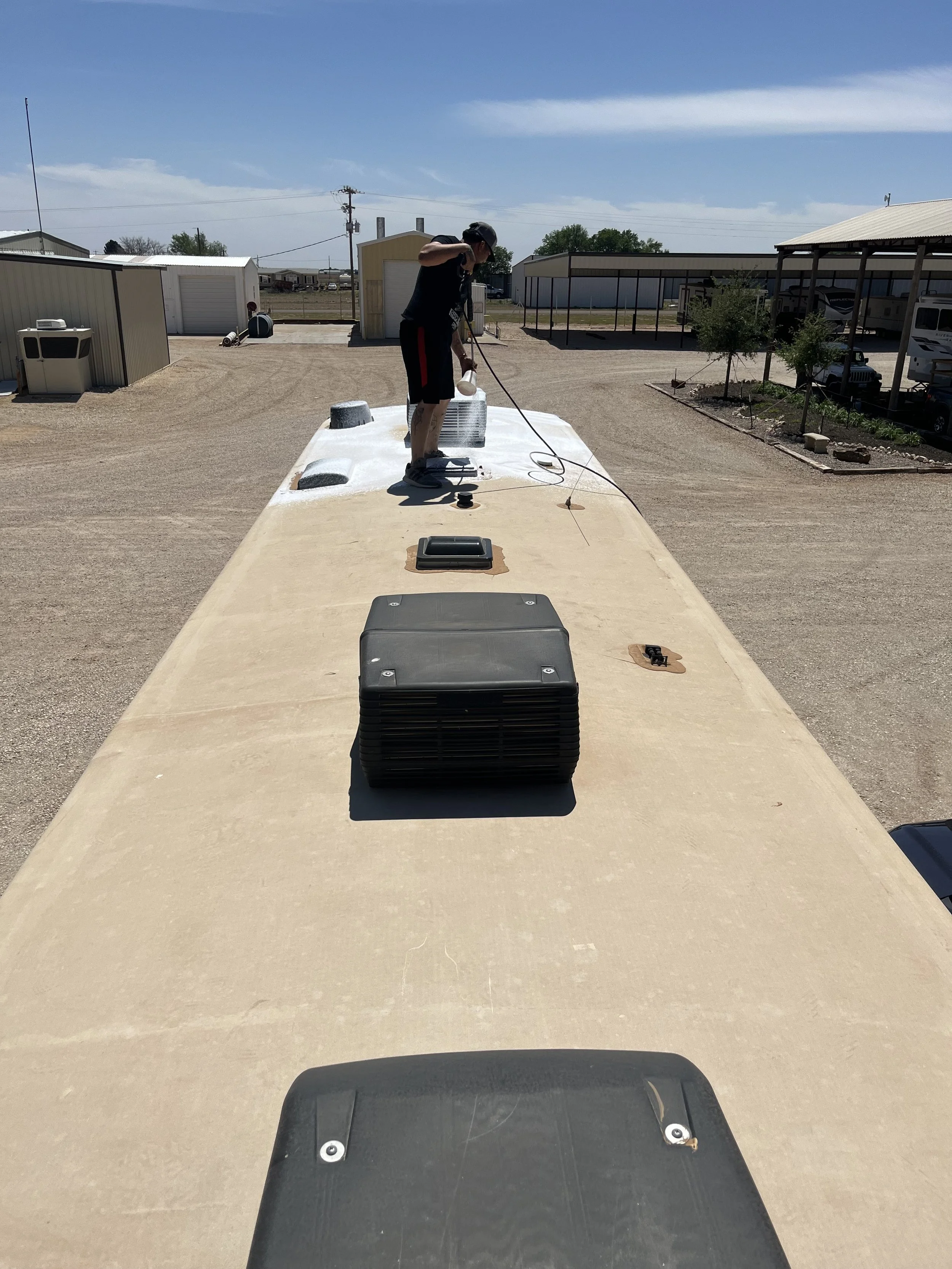 A person on top of a beige camper van is pressure washing the roof. The person is wearing black clothing and a black cap, and the camper is parked in a gravel lot with some buildings and trees in the background.