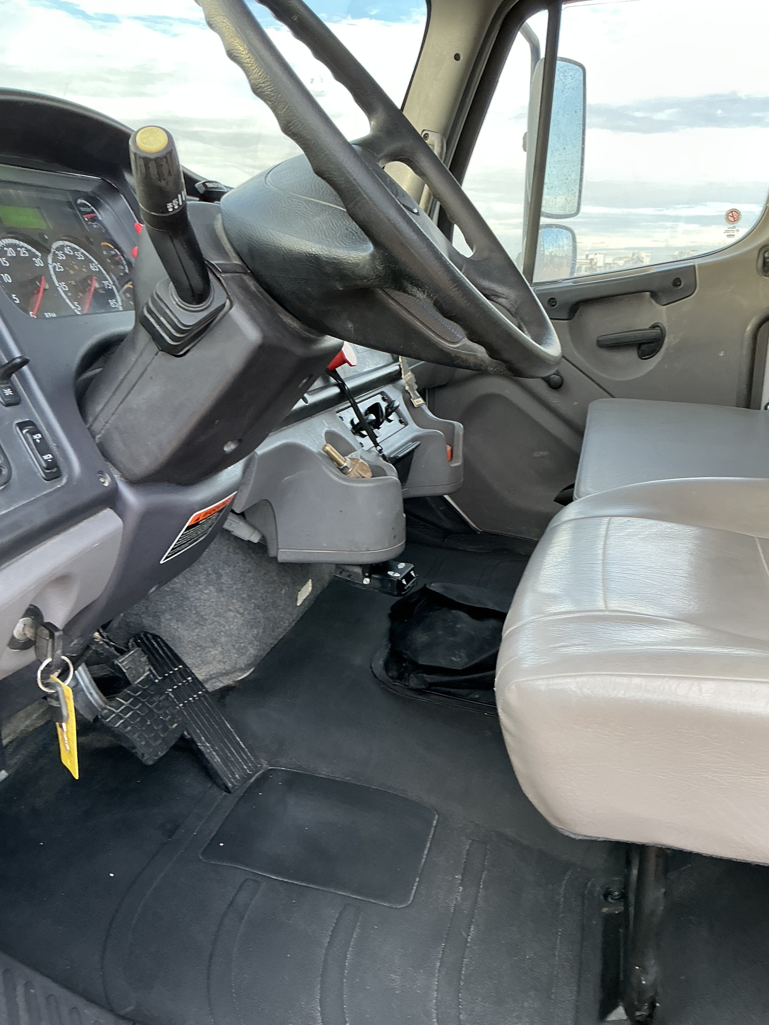 Inside of a truck cabin showing the steering wheel, dashboard, gear shift, driver’s seat, and the pedals on a black rubber floor mat.