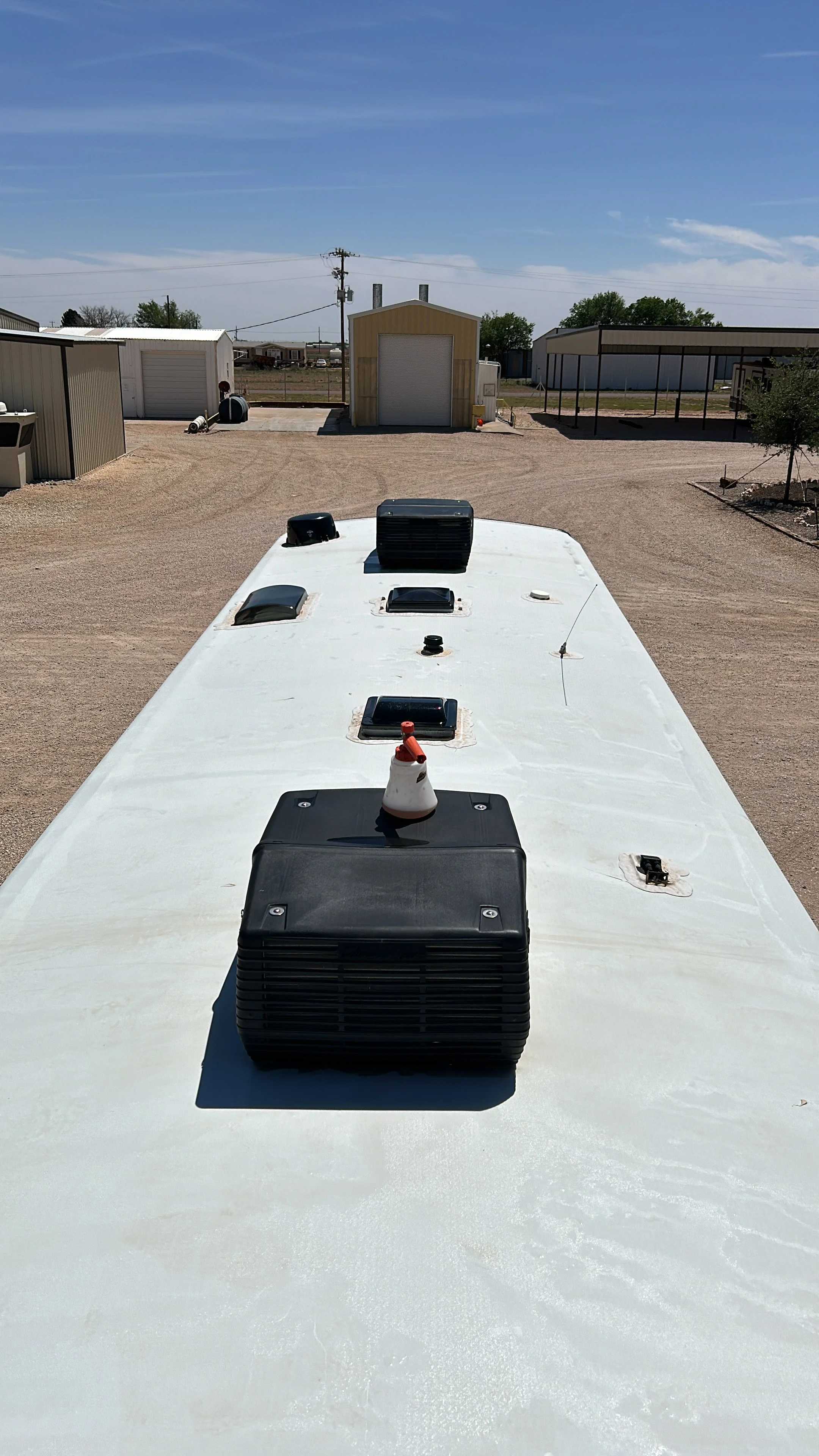 View from the roof of a travel trailer showing vents and an air conditioning unit, with storage buildings and a gravel lot in the background under a clear blue sky.