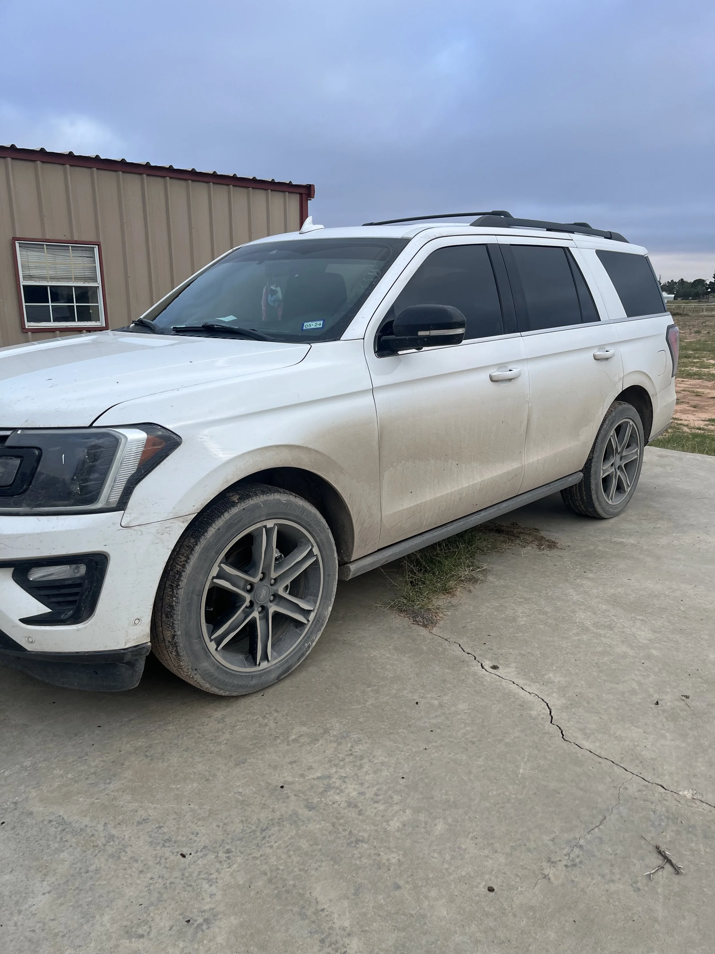 A white SUV parked on a concrete surface next to a beige metal building, with a cloudy sky overhead.