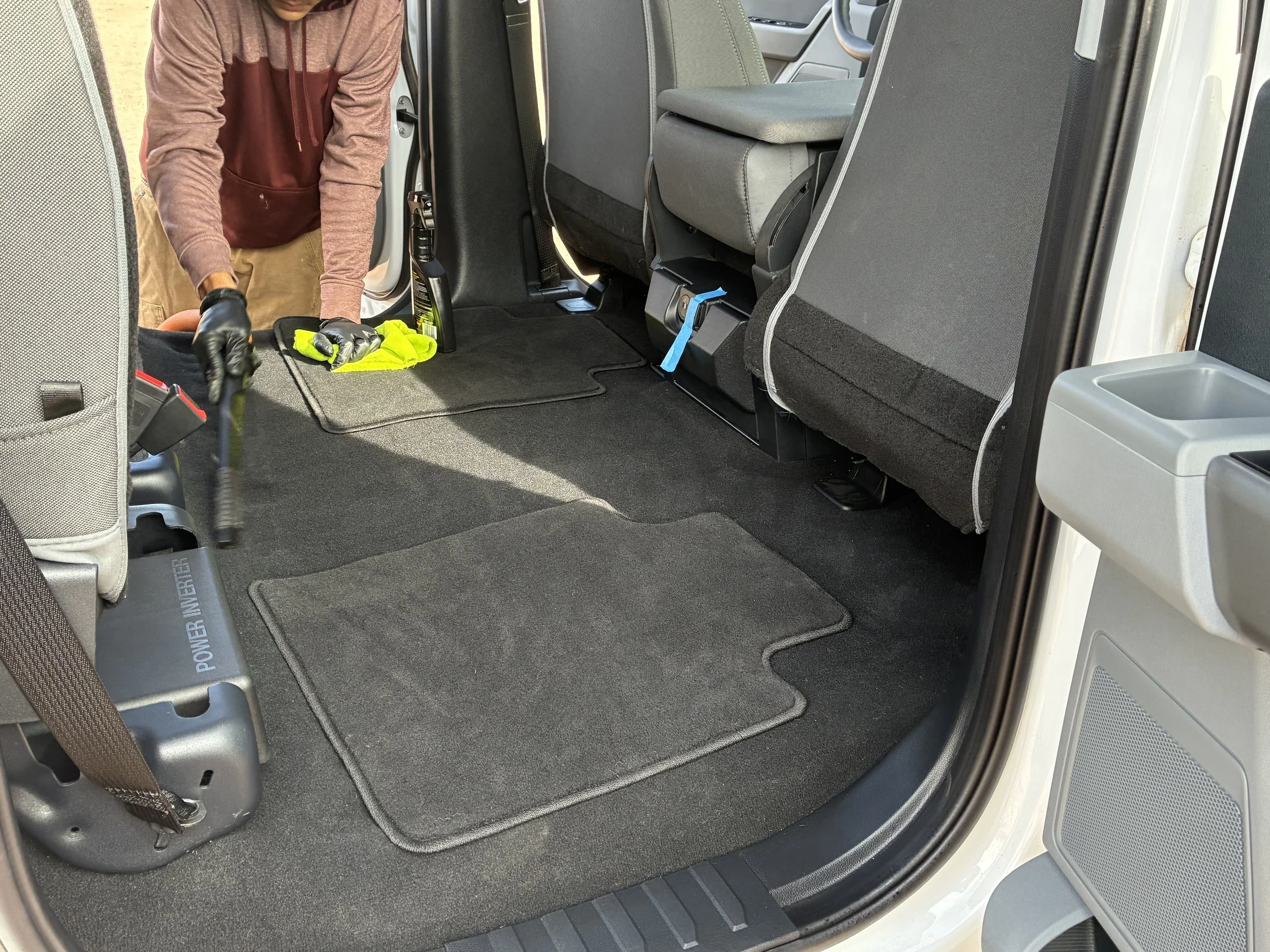 Person wearing gloves cleaning the interior of a vehicle, focusing on the carpeted floor mats and seats.