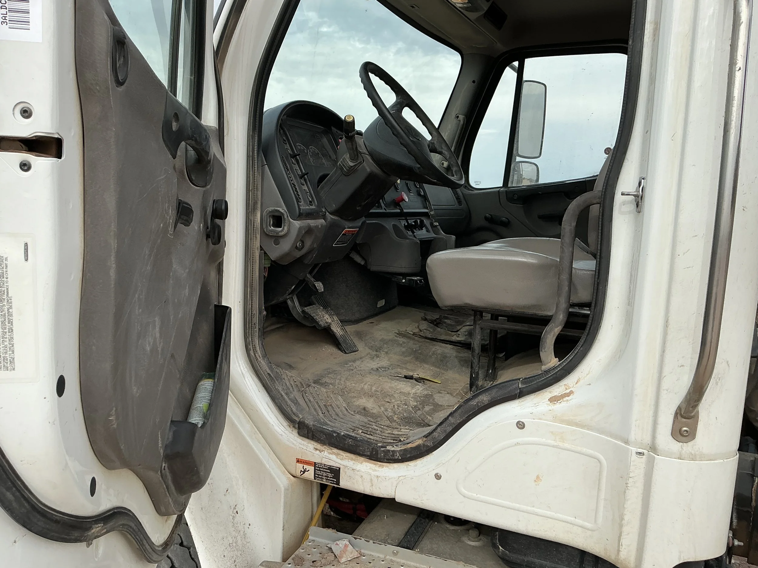 Interior of a dirty, worn construction vehicle cab with a steering wheel, dashboard, and gray seat, accessed via an open door.