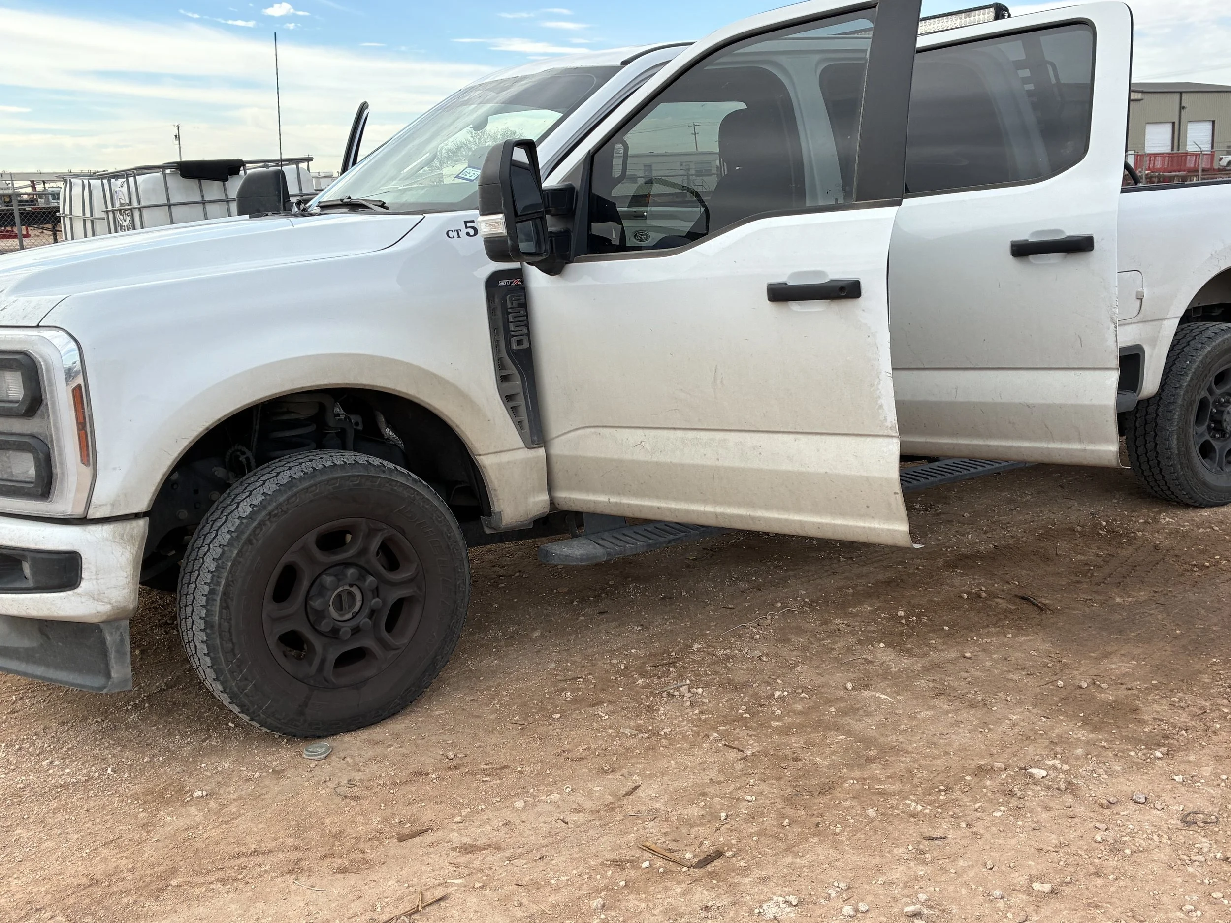 A white pickup truck with black wheels and side mirrors, parked on a dirt surface. The truck's front right tire is slightly turned, and the door on the driver's side is open.
