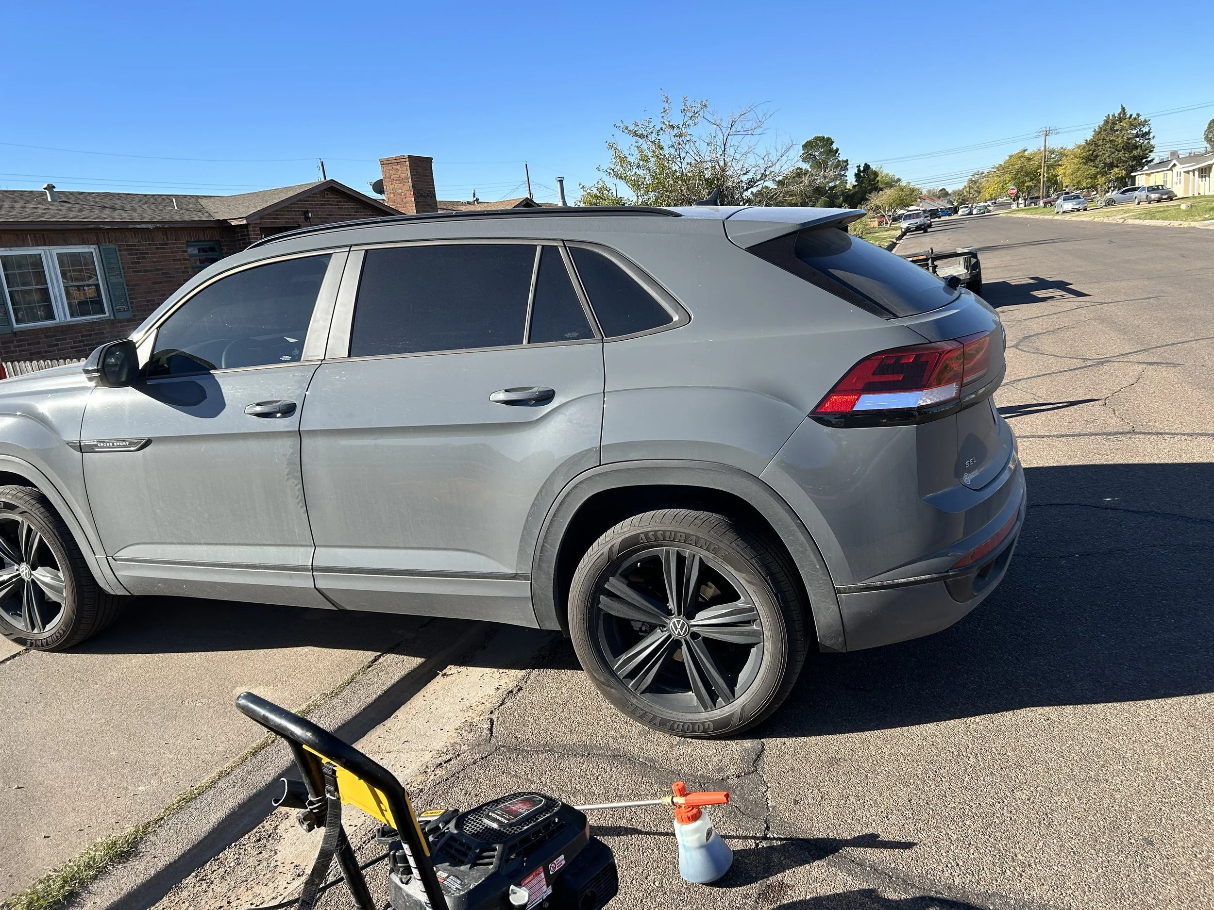 A grey SUV parked on a residential street under clear blue sky. There is a wheel cleaning tool and spray bottle on the ground near it.