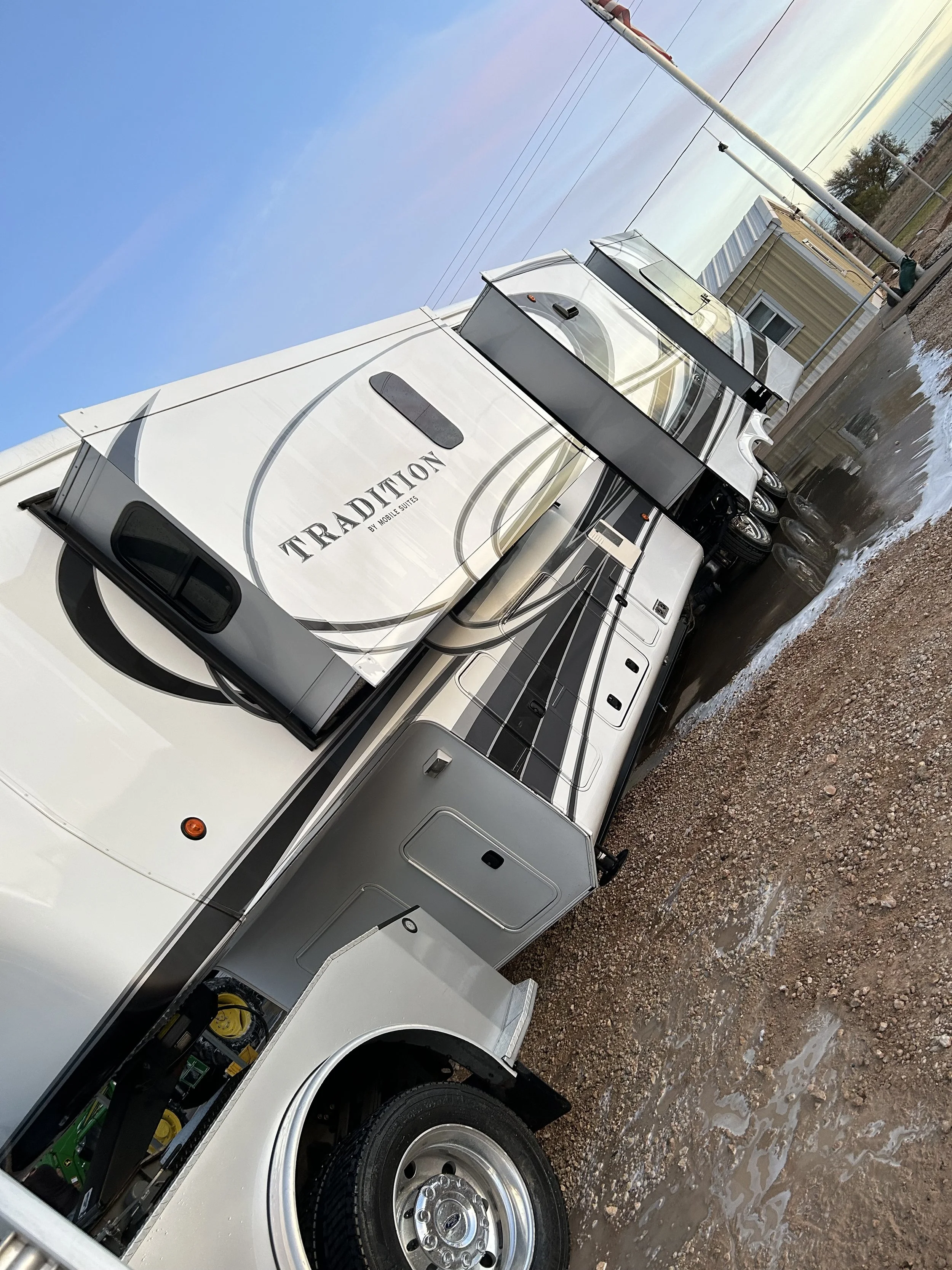 Travel trailer RV parked on a gravel lot with a yellow building and power lines in the background.