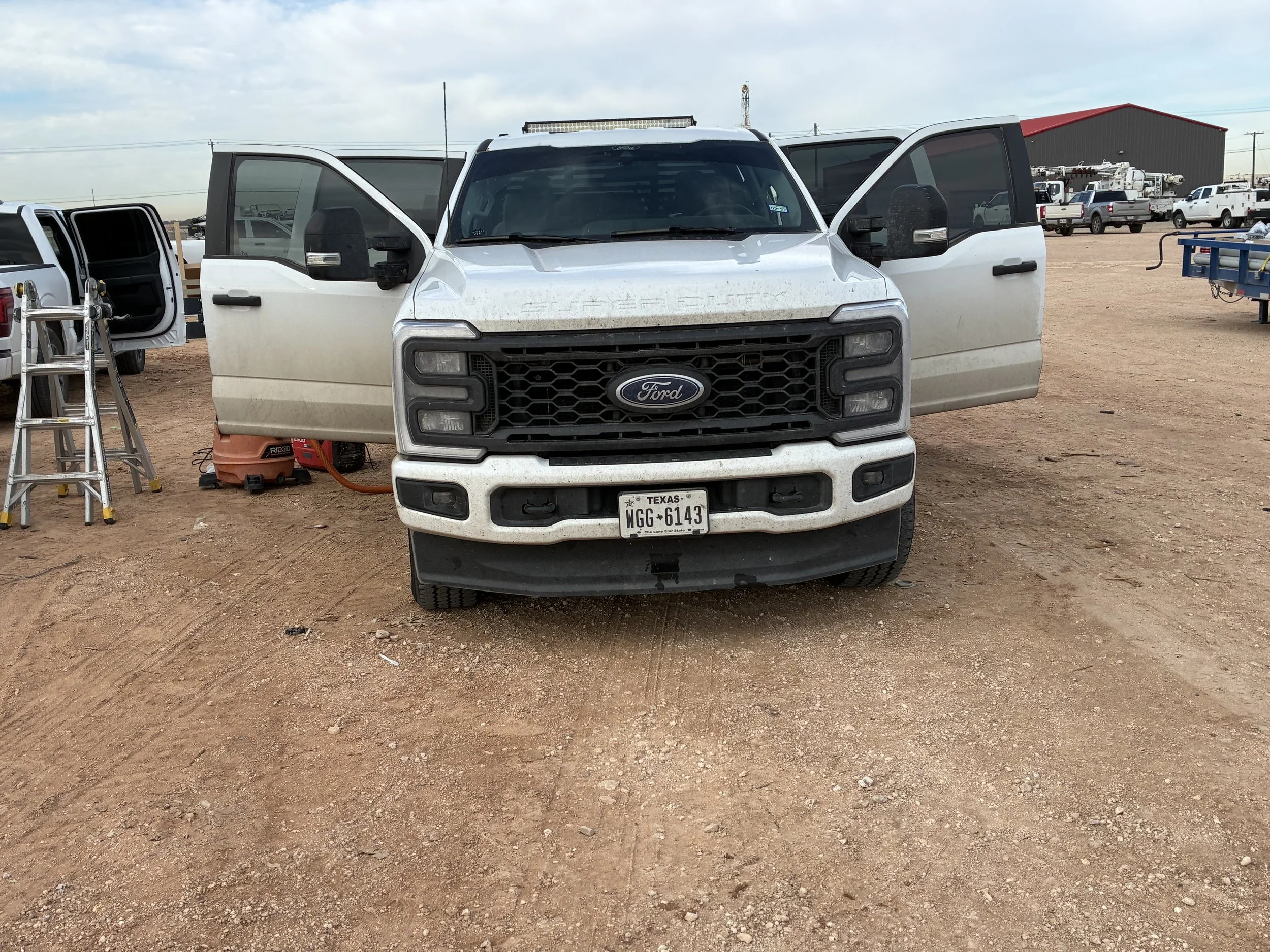 A white Ford pickup truck with open doors, parked on a dirt lot, surrounded by other vehicles and equipment.