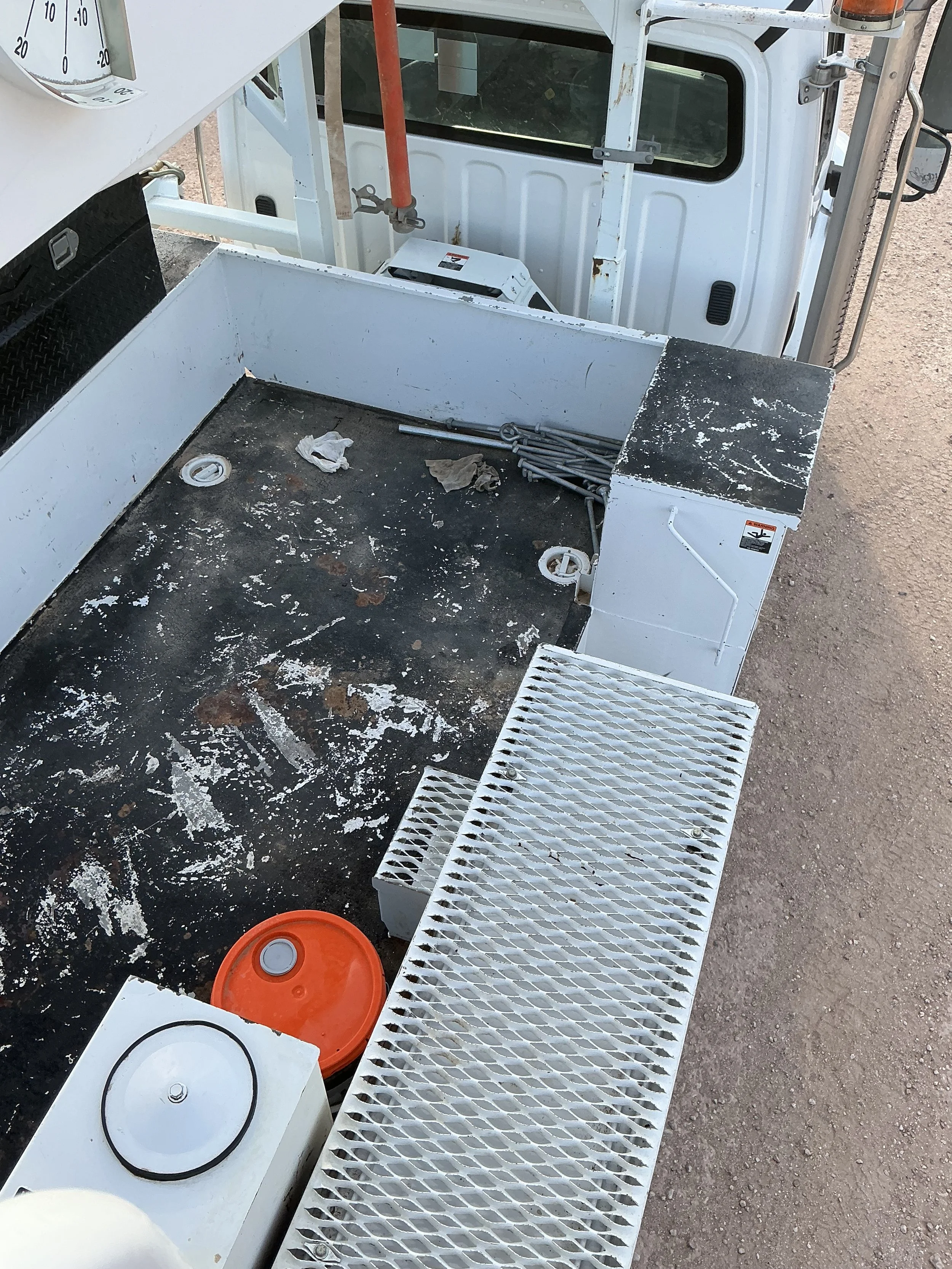 Utility bucket truck platform with tools, a red bucket, and a white grate, parked on a dirt surface.