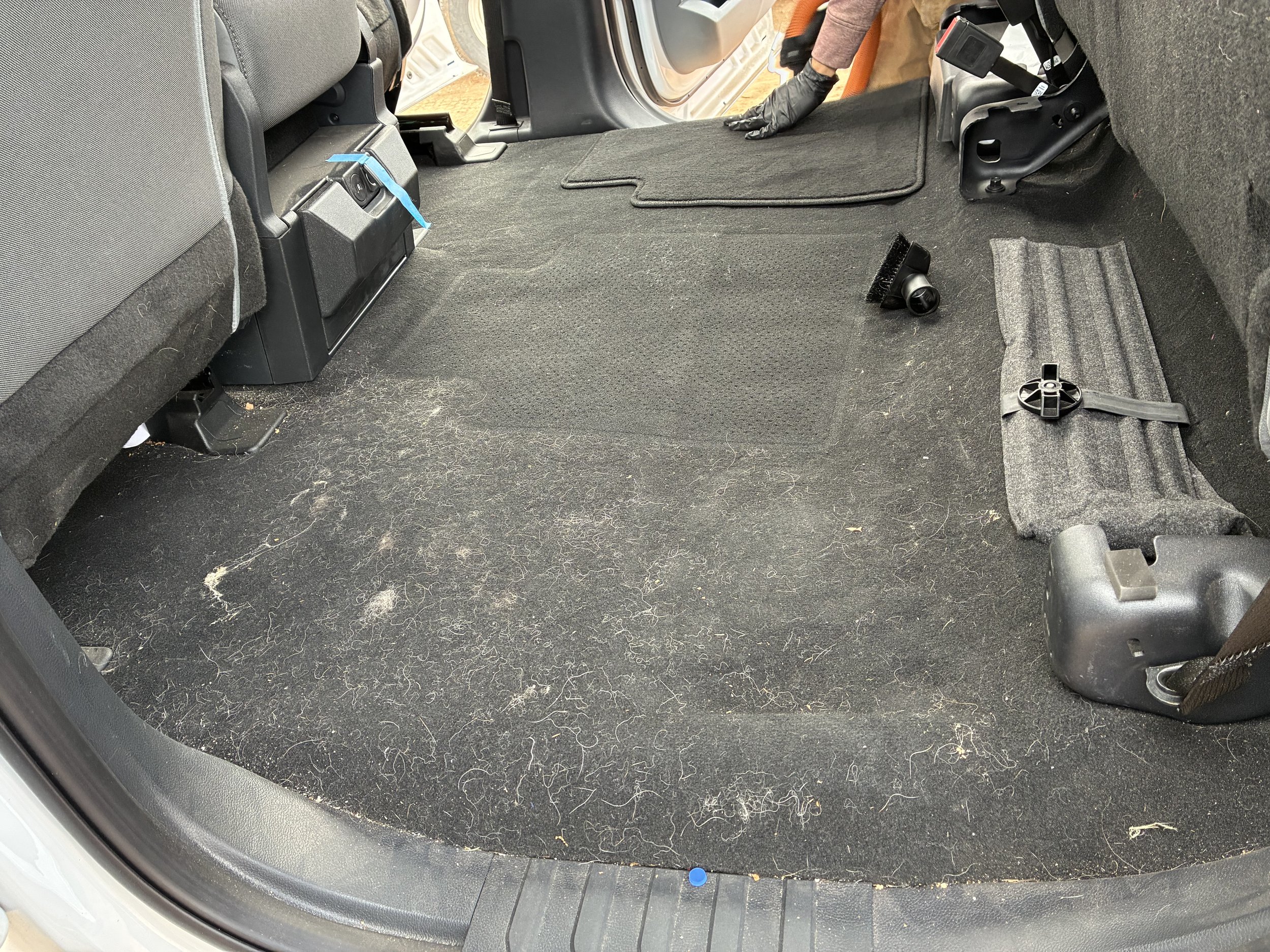 The back interior of a vehicle with the rear seats folded down, showing the black carpeted floor which is dusty and has hair and debris scattered across it. There are various tools and parts on the right side of the floor, and a person's gloved hand 