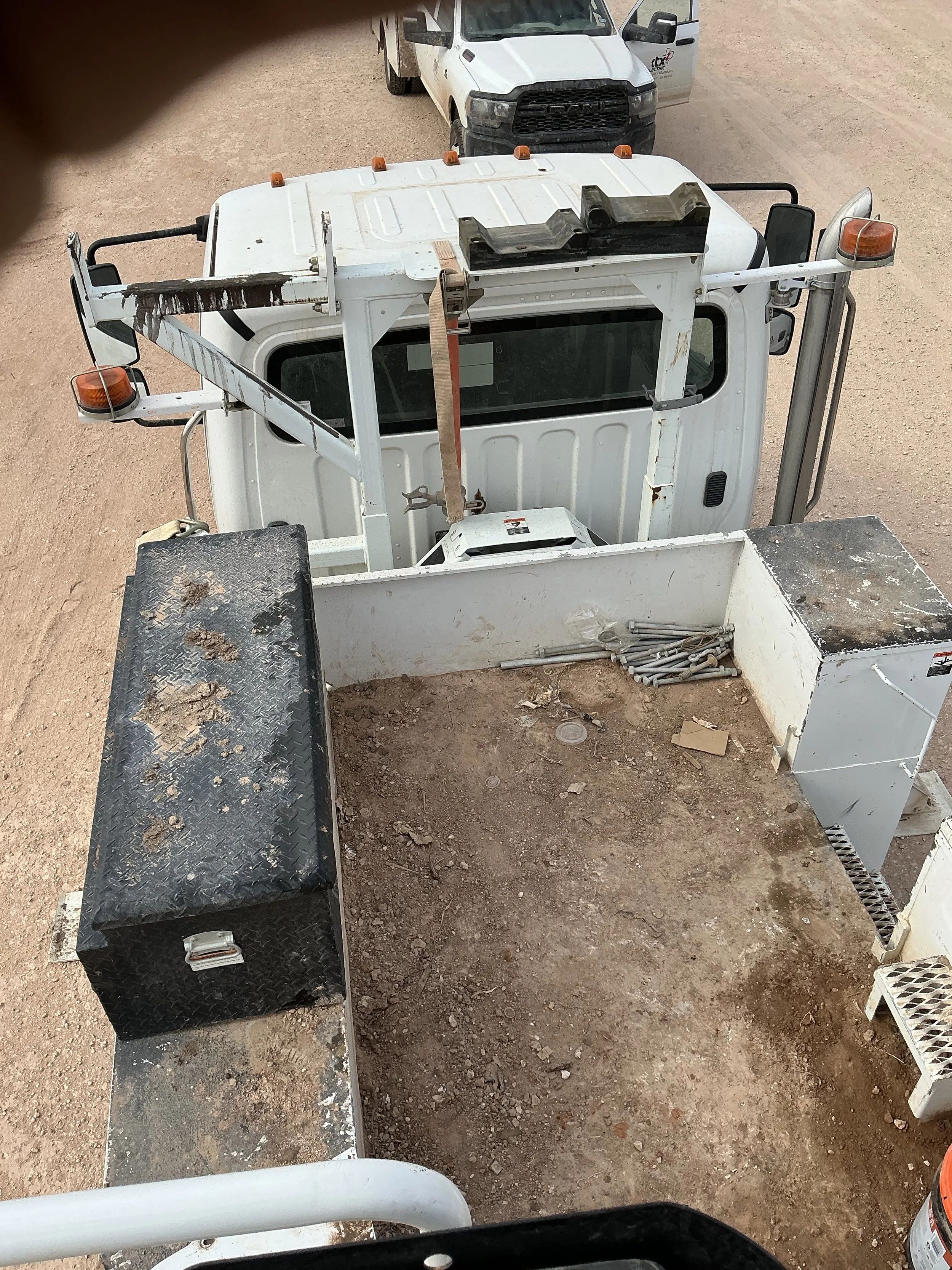 View from inside a utility or construction vehicle with a muddy and dirt-covered platform, black toolbox on the left, and various cables on the right, with other trucks visible in the background on a dirt surface.
