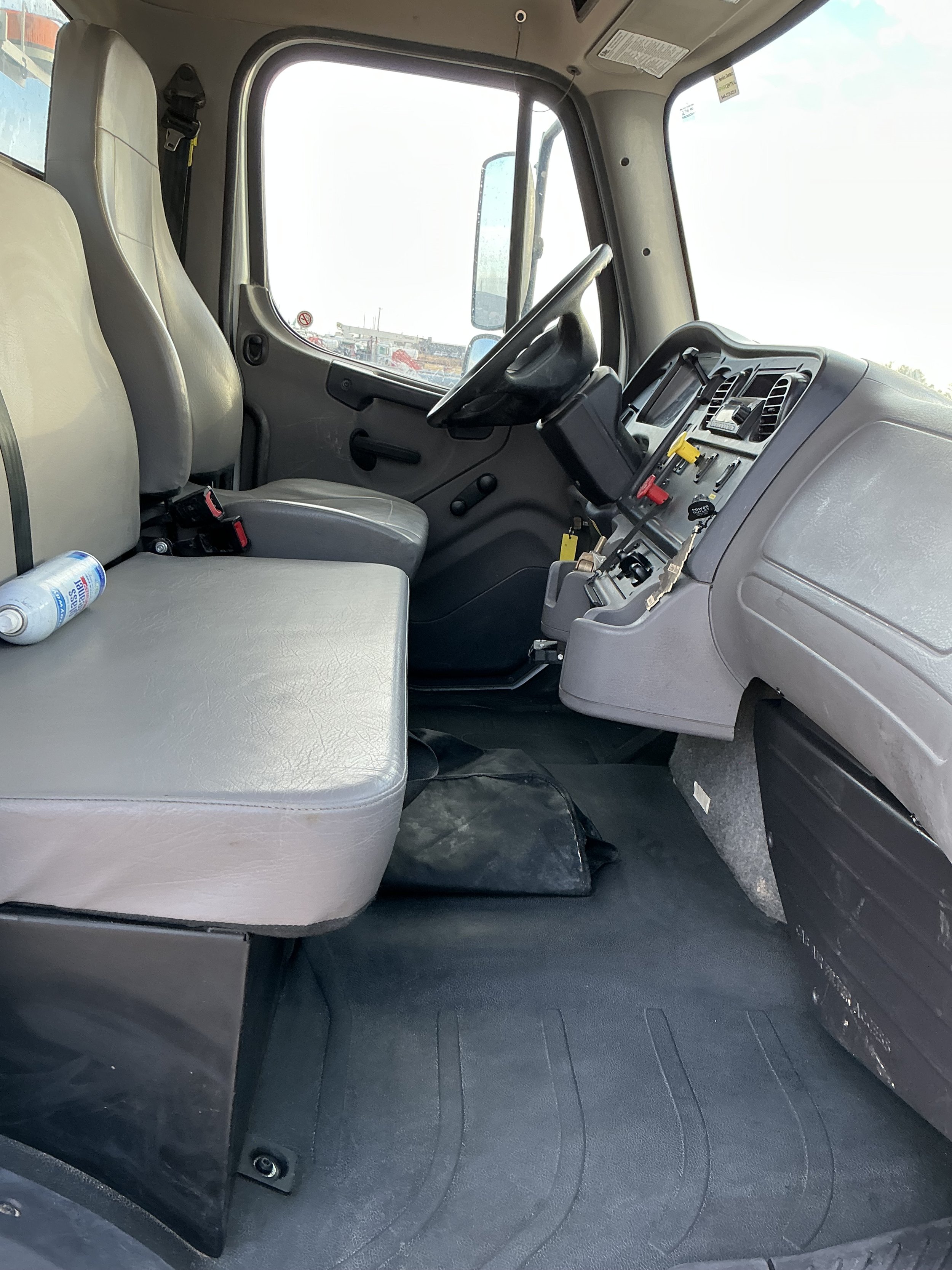 Interior of a truck cab, showing the driver's seat, passenger seat, steering wheel, dashboard with control panel, and a water bottle on the passenger seat.