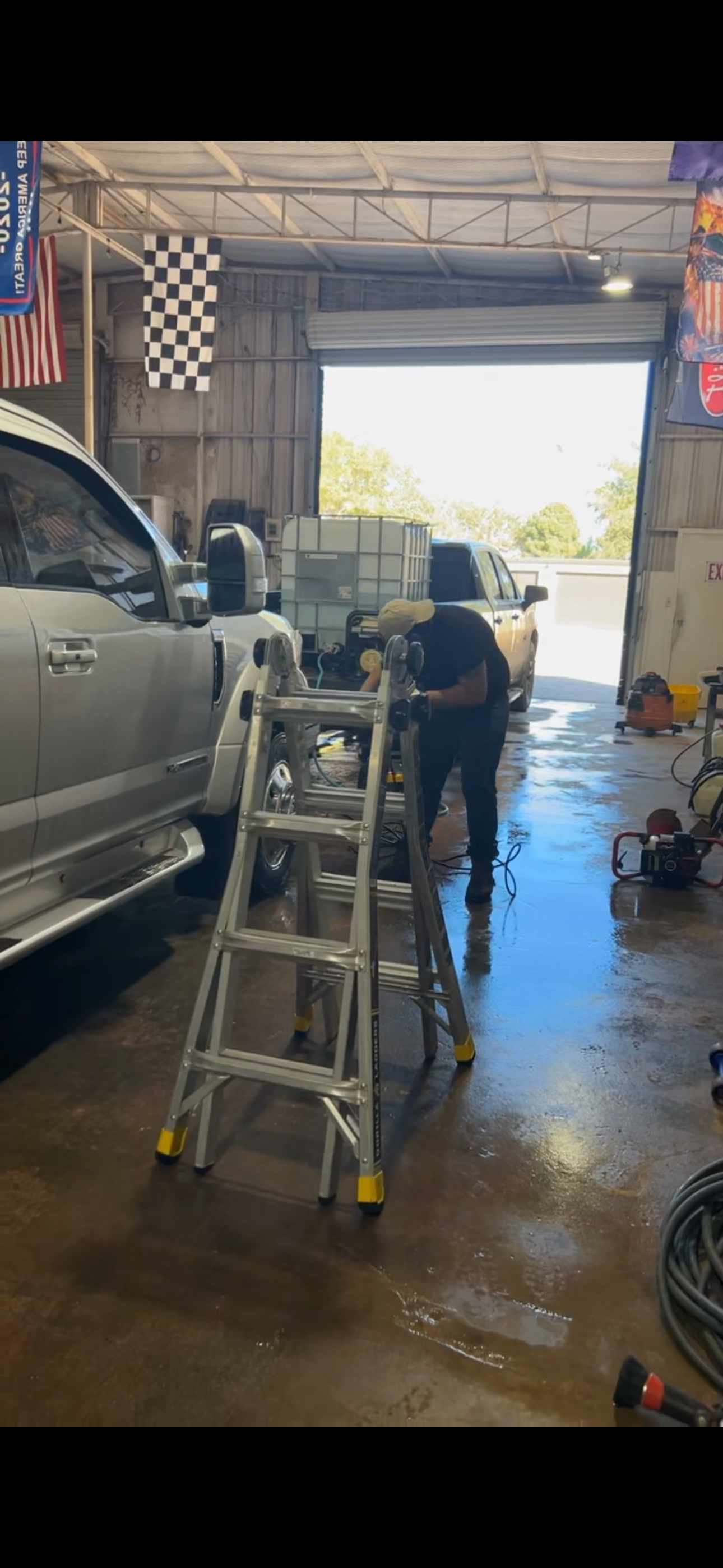 A person working on a silver pickup truck inside a garage with an open roll-up door, mountains of water containers outside, and various tools and equipment scattered around.