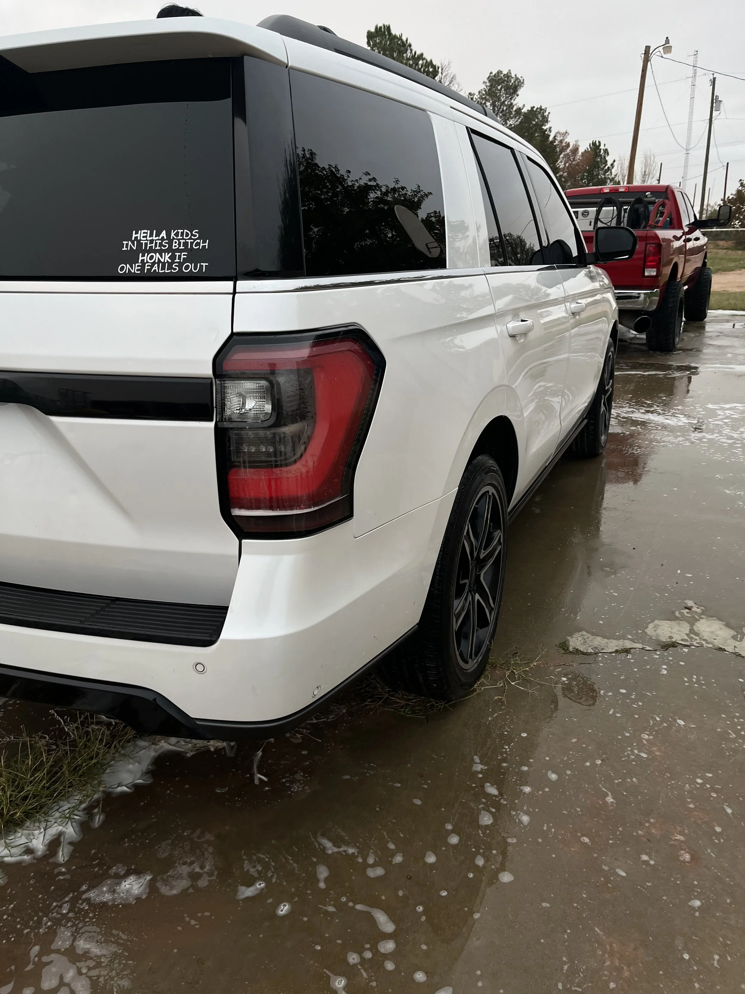 White vehicle with a black sticker on the rear window that reads: "HELLA KIDS IN THIS BITCH HONK IF ONE FALLS OUT"; parked on a wet surface with a red truck visible in the background.